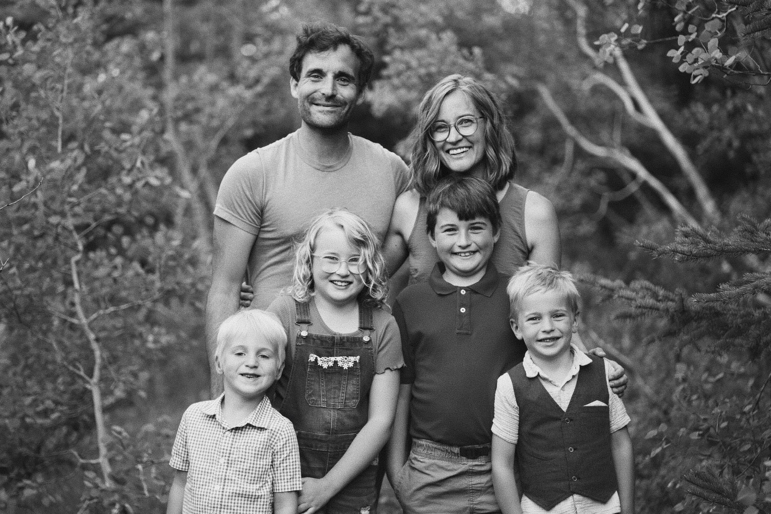 Black-and-white portrait of a family of six smiling together among the trees, the four children gathered in front of their parents.