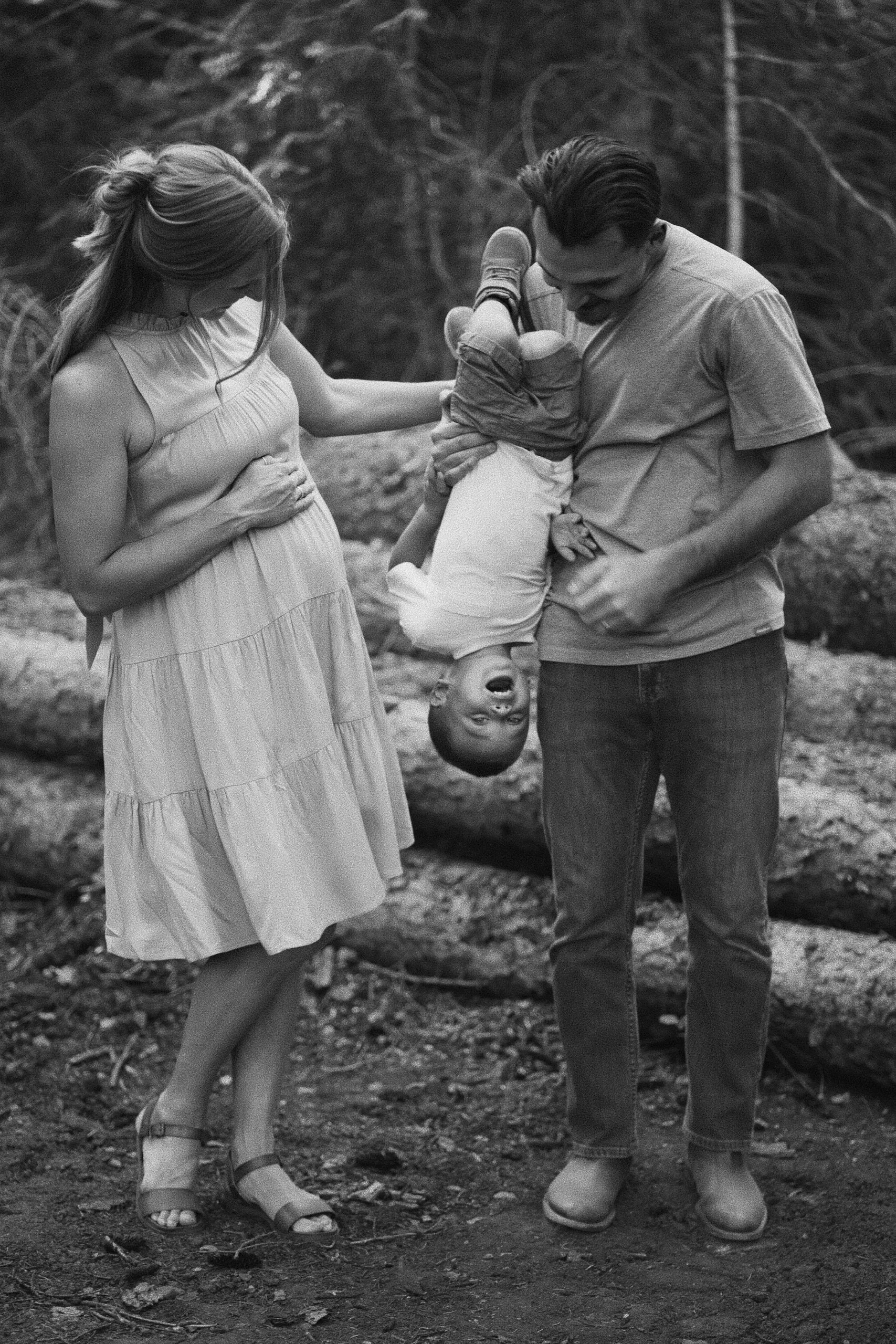 Black-and-white photo of an expecting mother in a dress and a father playfully holding their young child upside down between them in the woods.