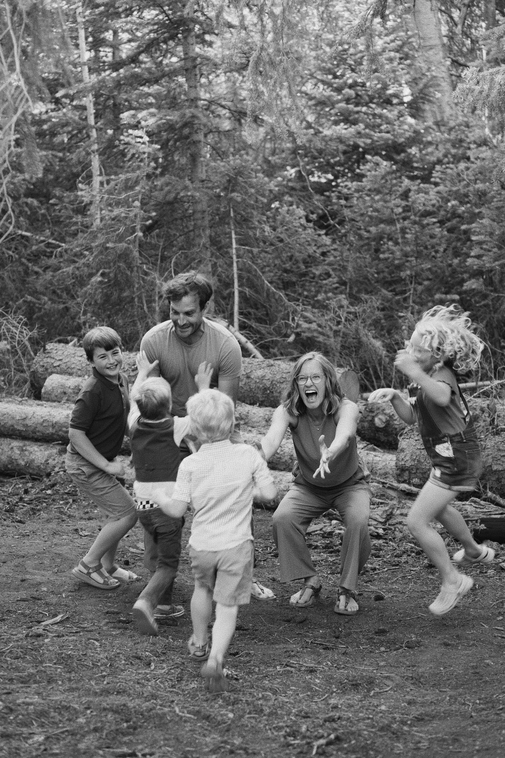 Black-and-white photo of a family laughing and playing in a circle in the forest, the children and parents mid-motion.