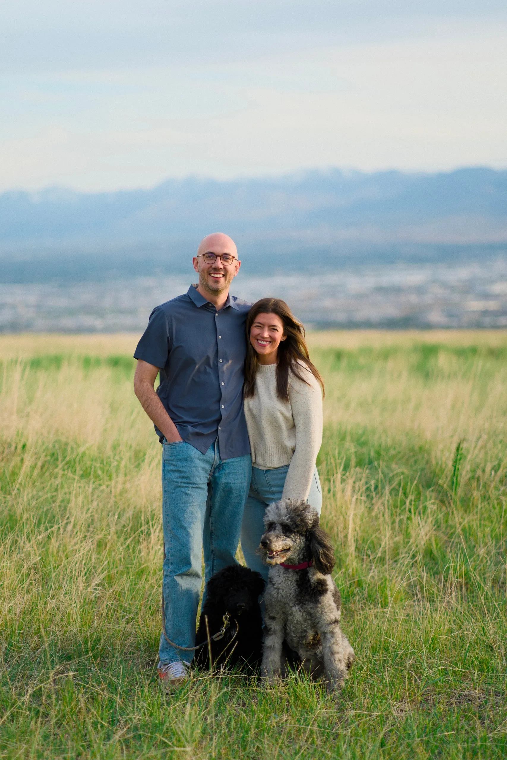 A couple embraces on a grassy hilltop above the Salt Lake valley at golden hour, their gray poodle sitting at their feet.