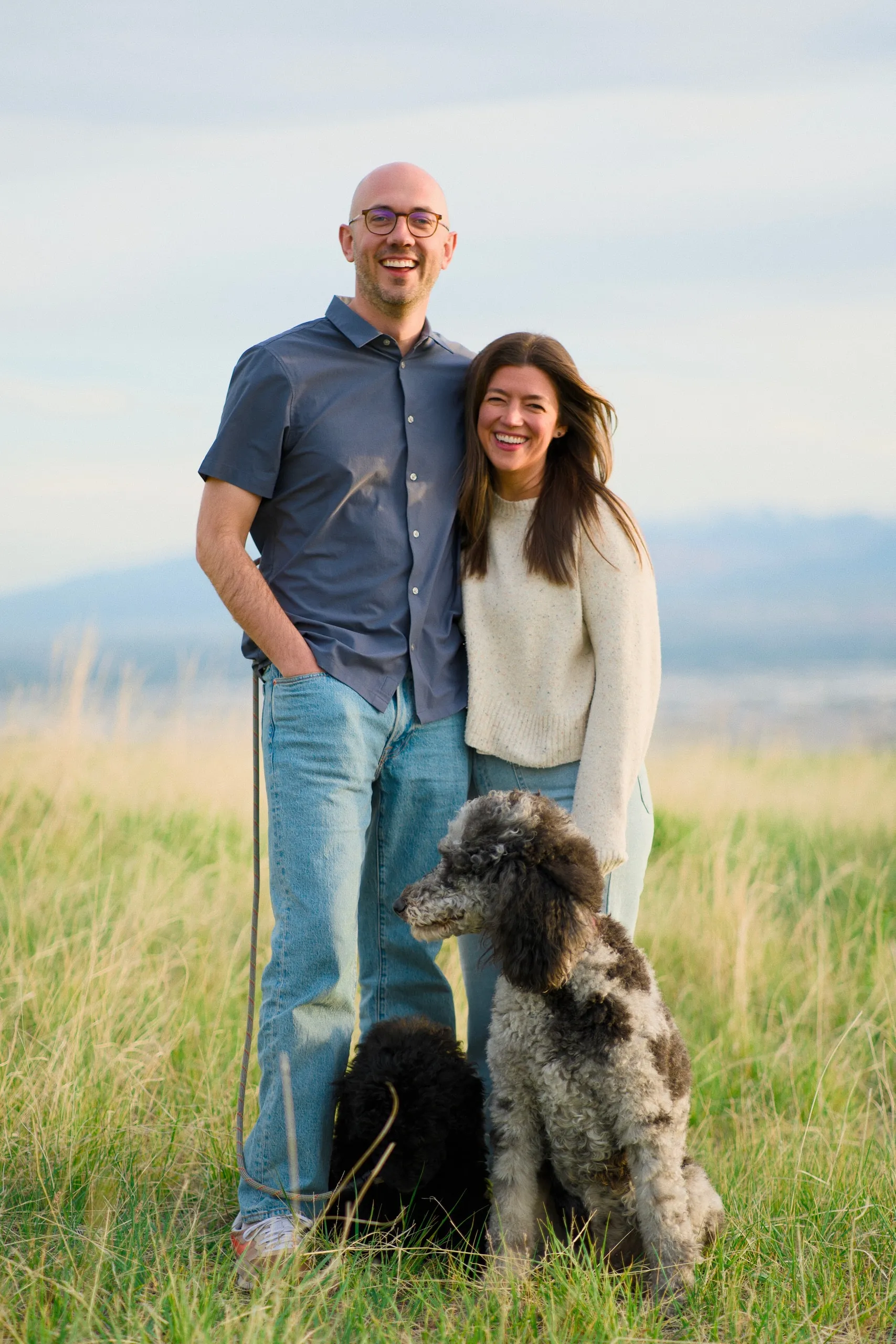 A smiling couple stand together on a grassy ridge overlooking the valley, with their gray poodle in front of them.