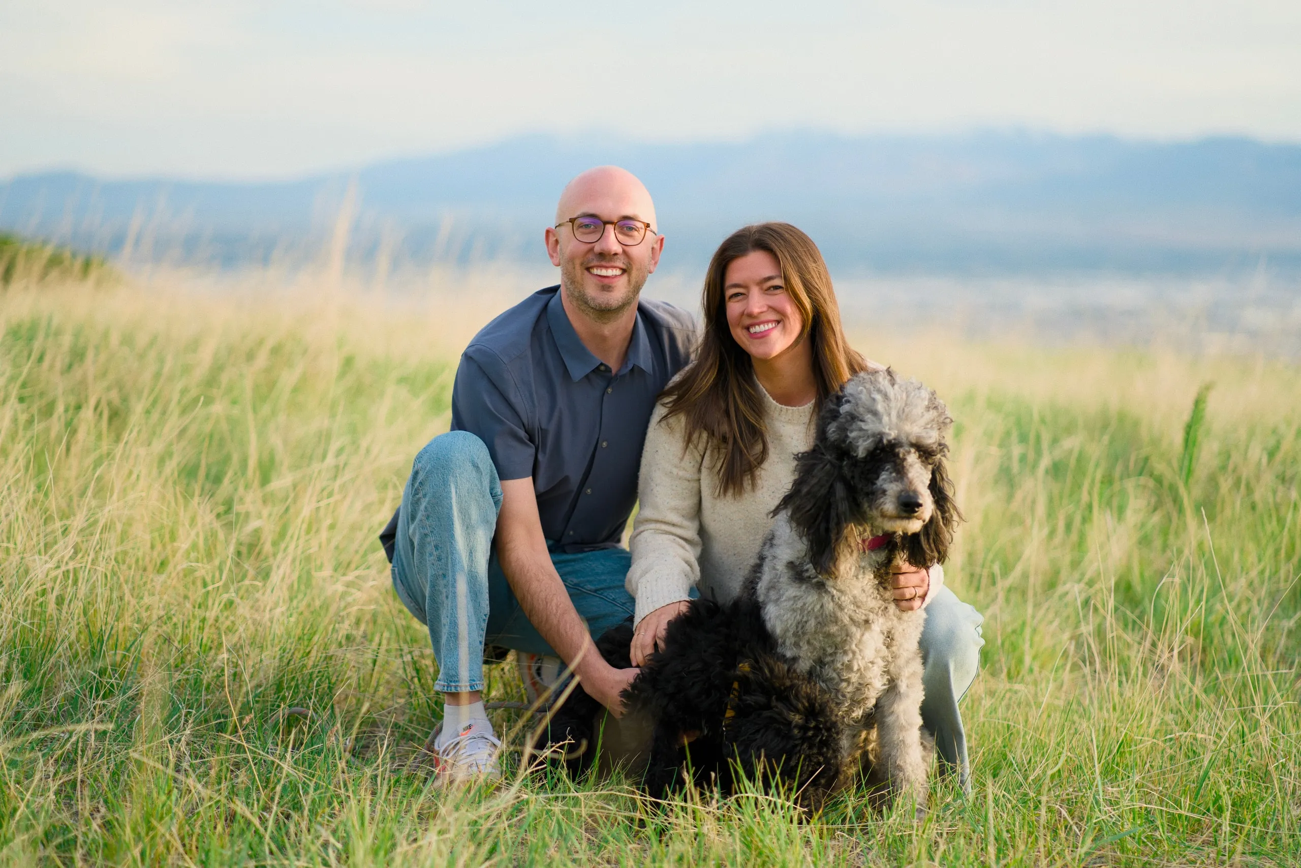 A couple crouch in a golden field with their arms around their shaggy gray-and-black dog, mountains in the distance.