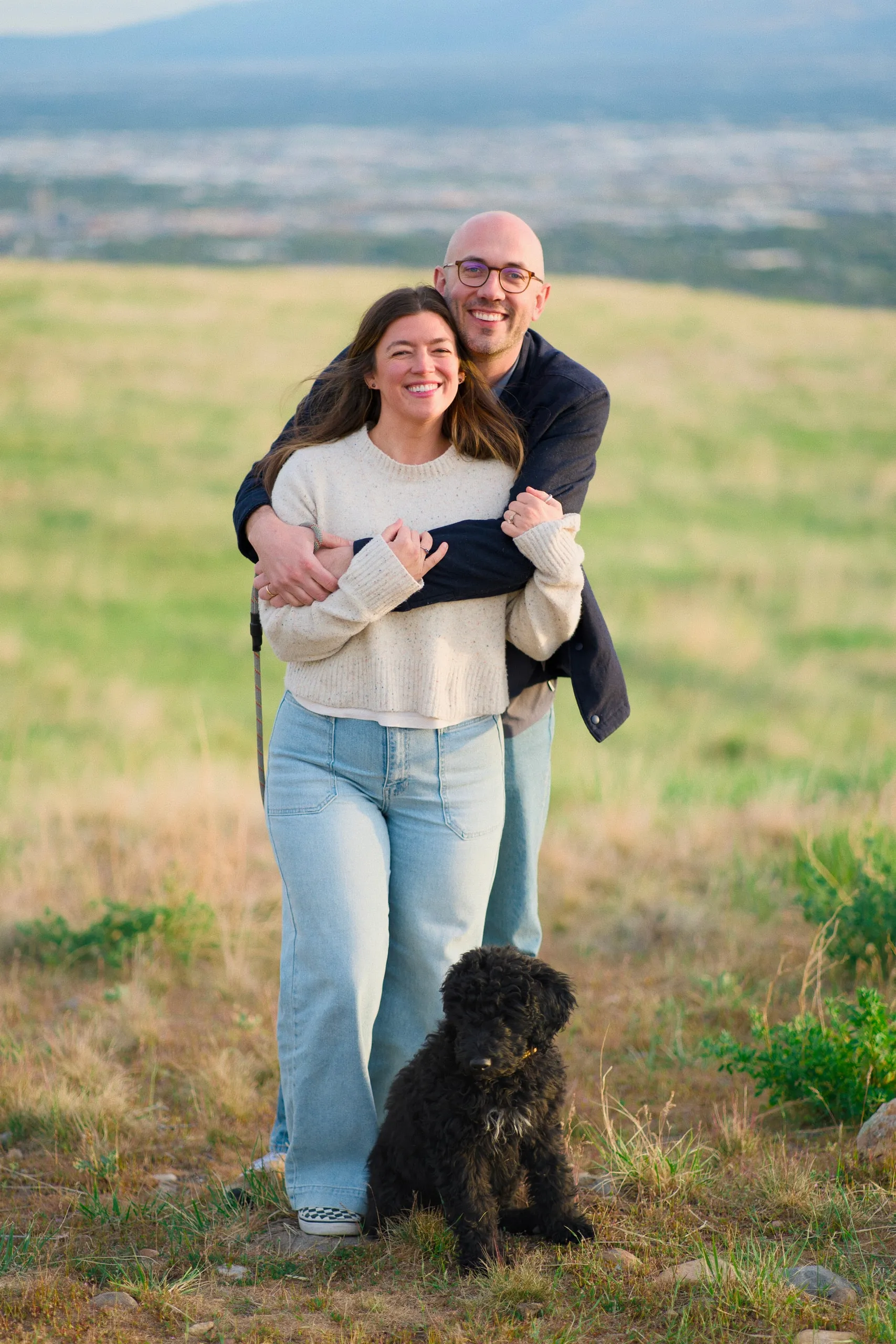 A man hugs a laughing woman from behind on a hilltop overlooking the valley, their small black dog sitting in the grass.