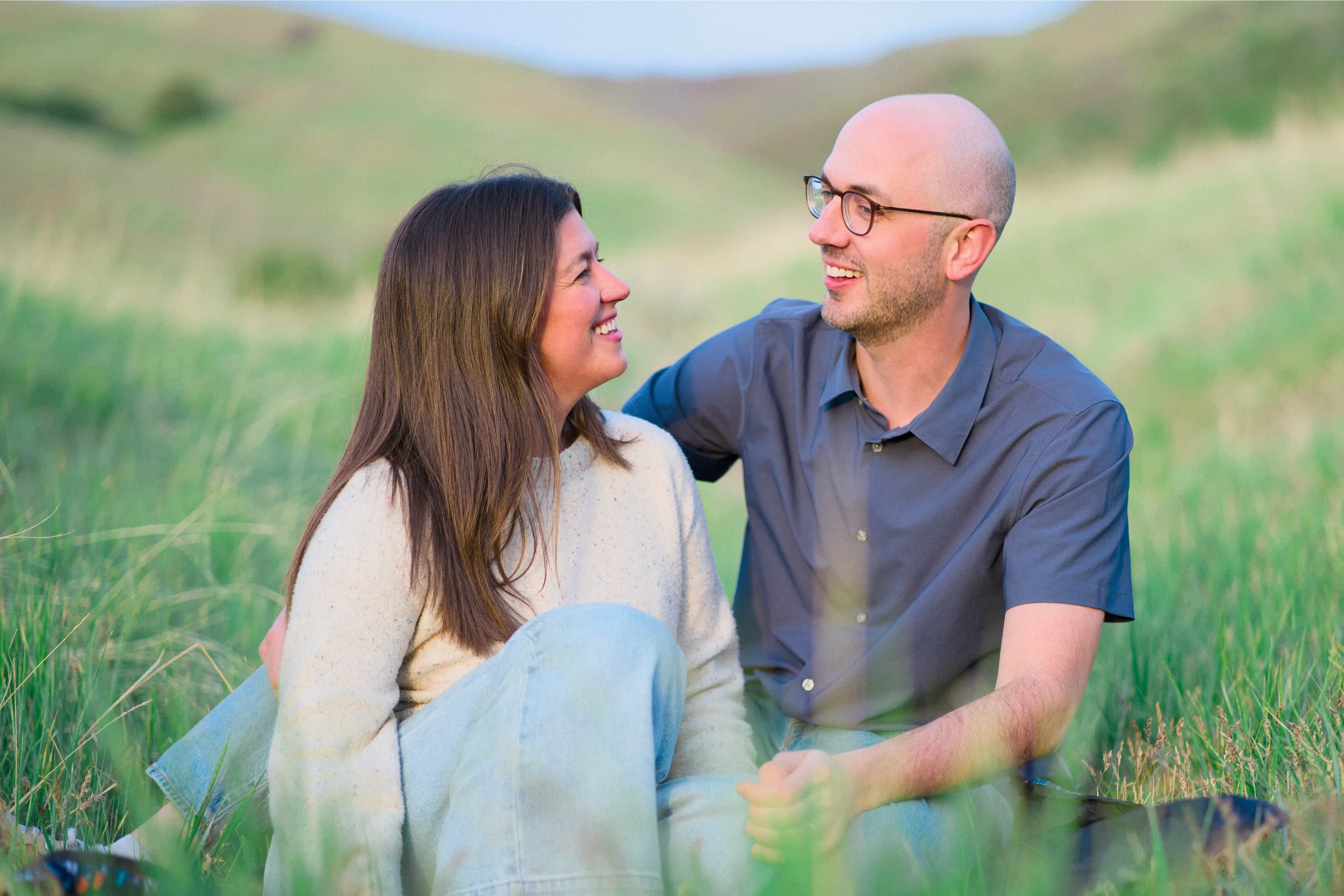 A couple sit together in tall grass at dusk, smiling at each other.