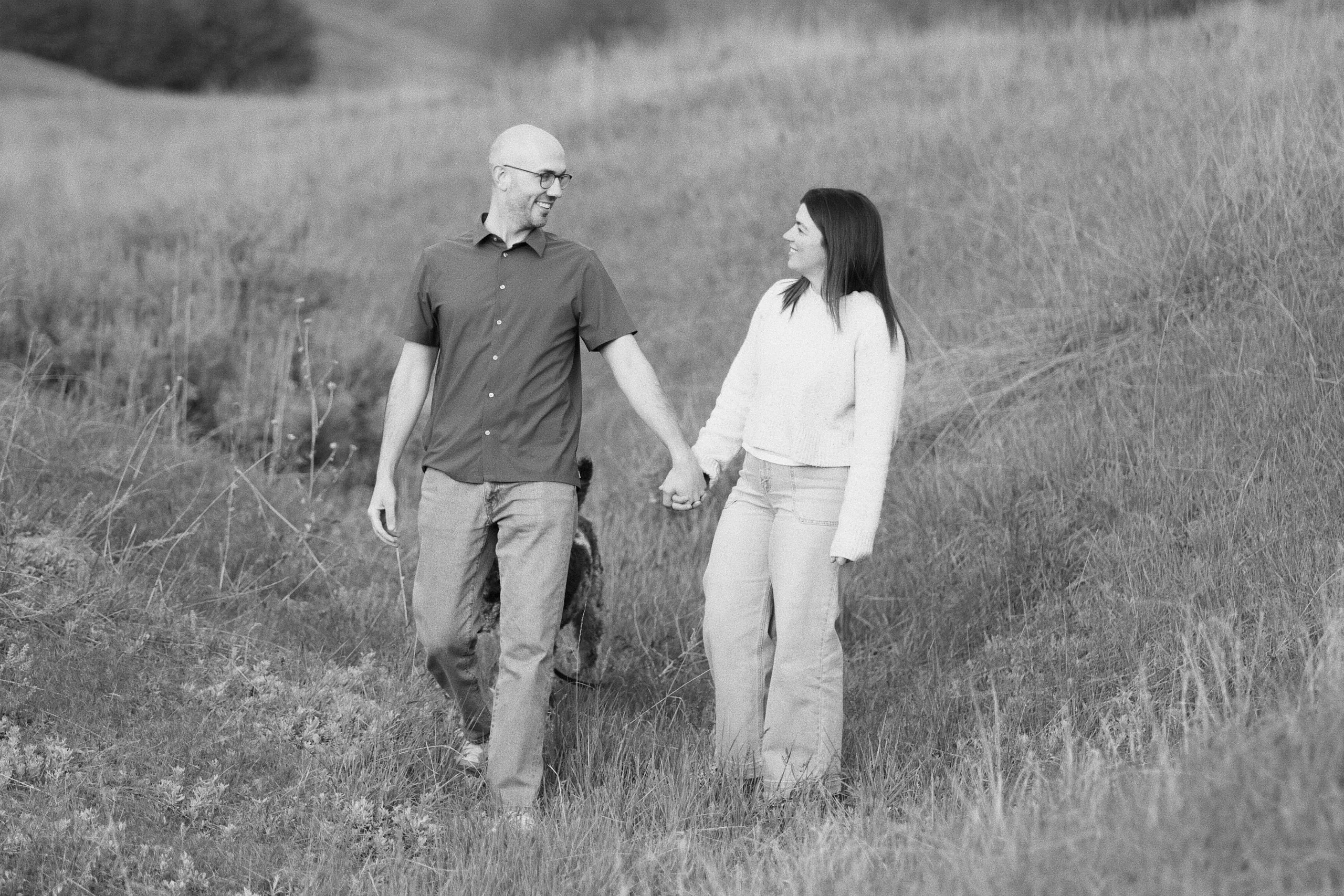 Black-and-white photo of a couple holding hands as they walk down a grassy hillside, looking at one another.
