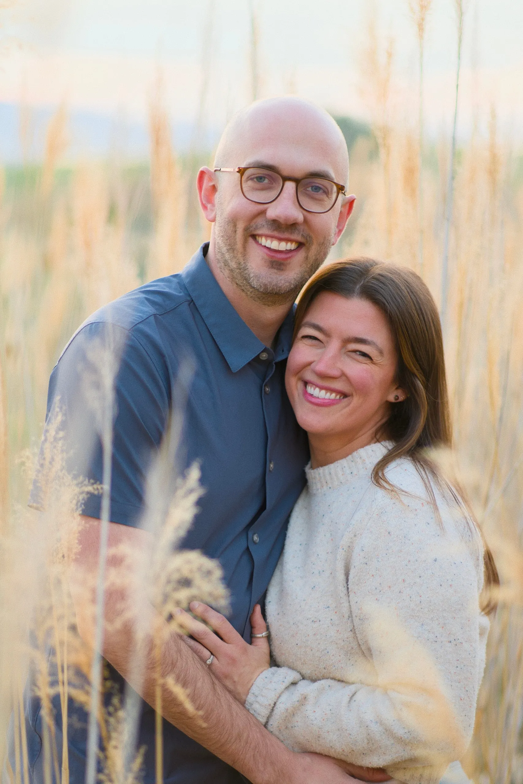 A couple embrace among tall golden grasses at sunset, smiling at the camera.