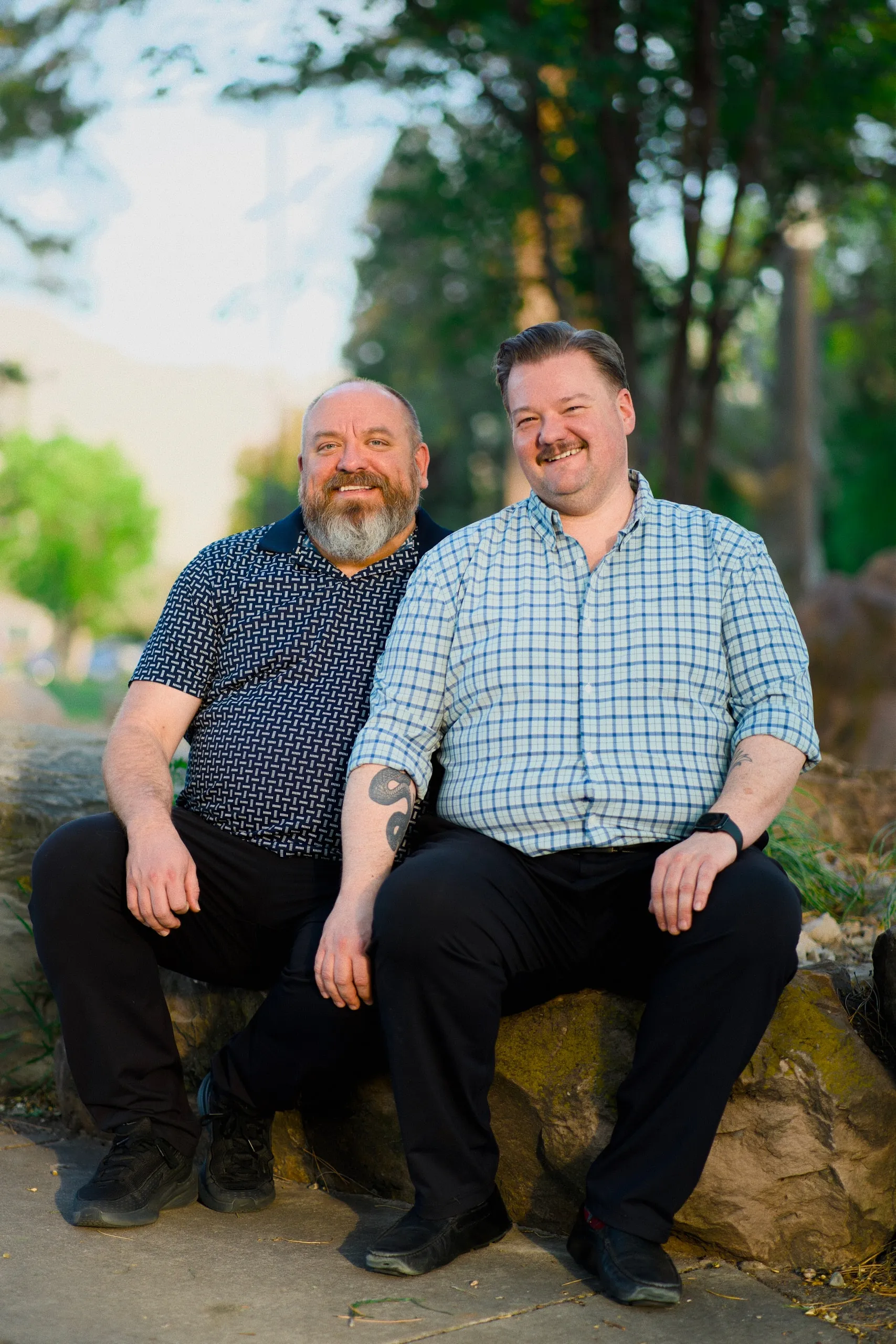 Two men sit together on a low stone wall in a park, smiling at the camera.