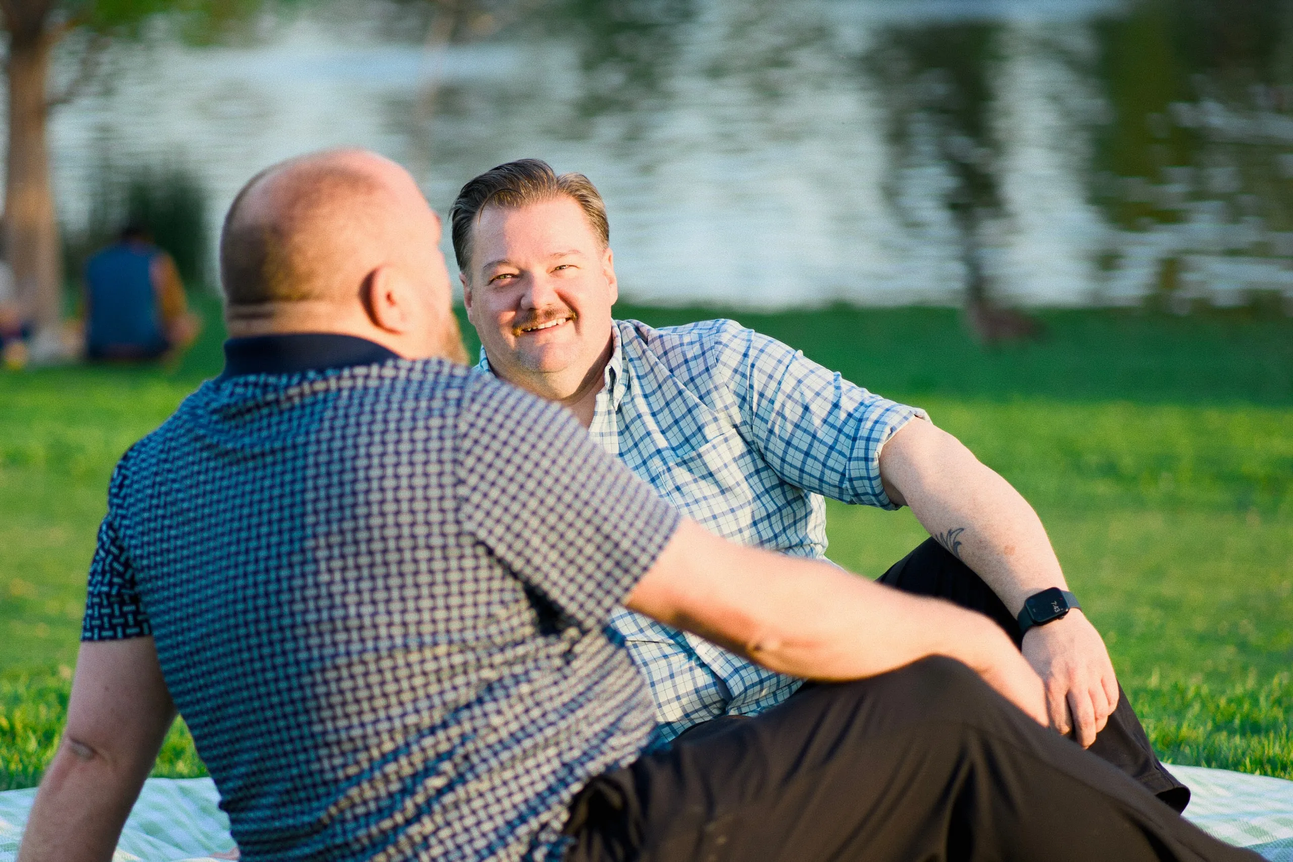 Two men sit on the grass by a lake at golden hour, laughing together.