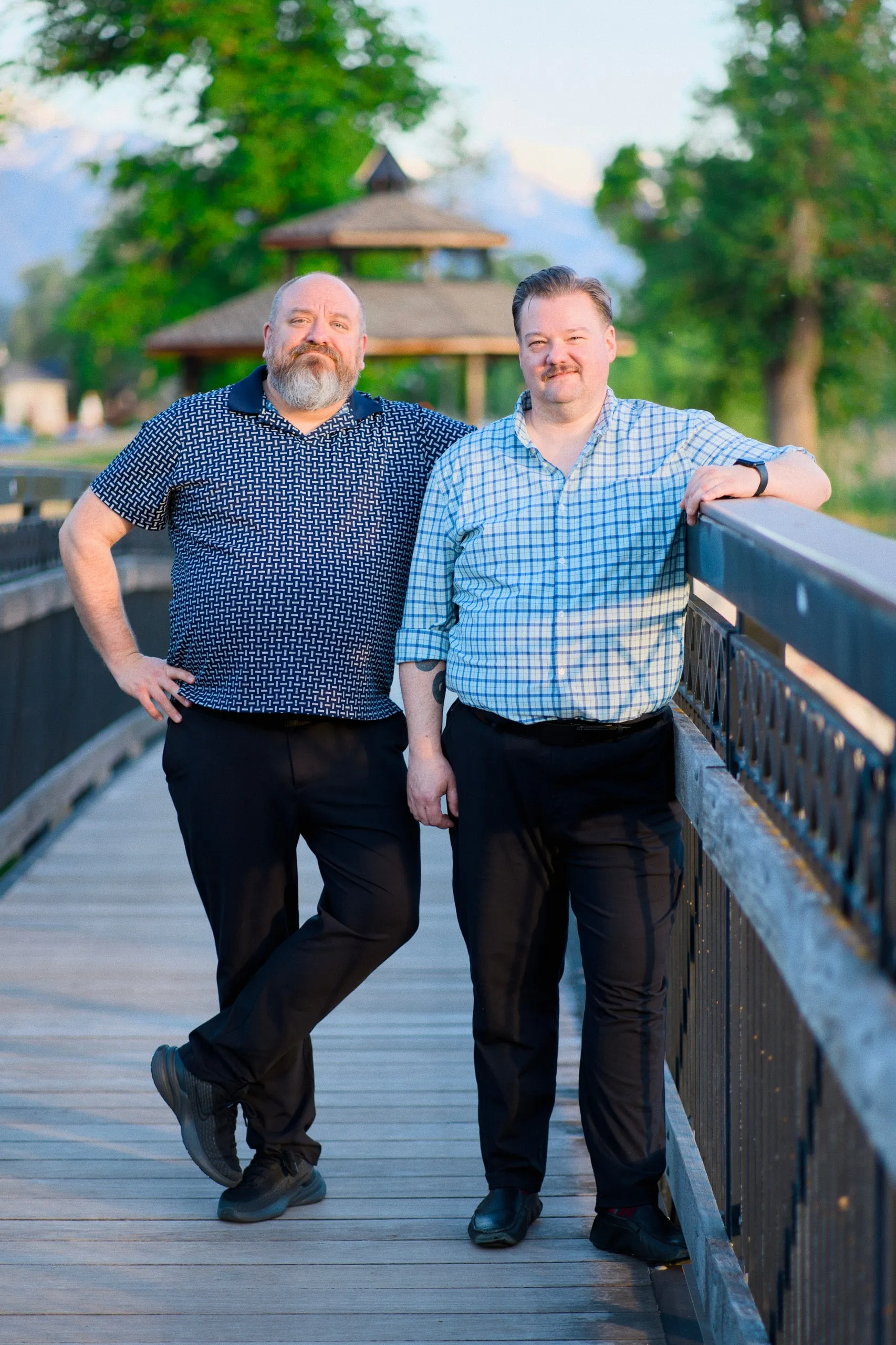 Two men lean against the railing of a wooden footbridge, with mountains and a park pavilion behind them.