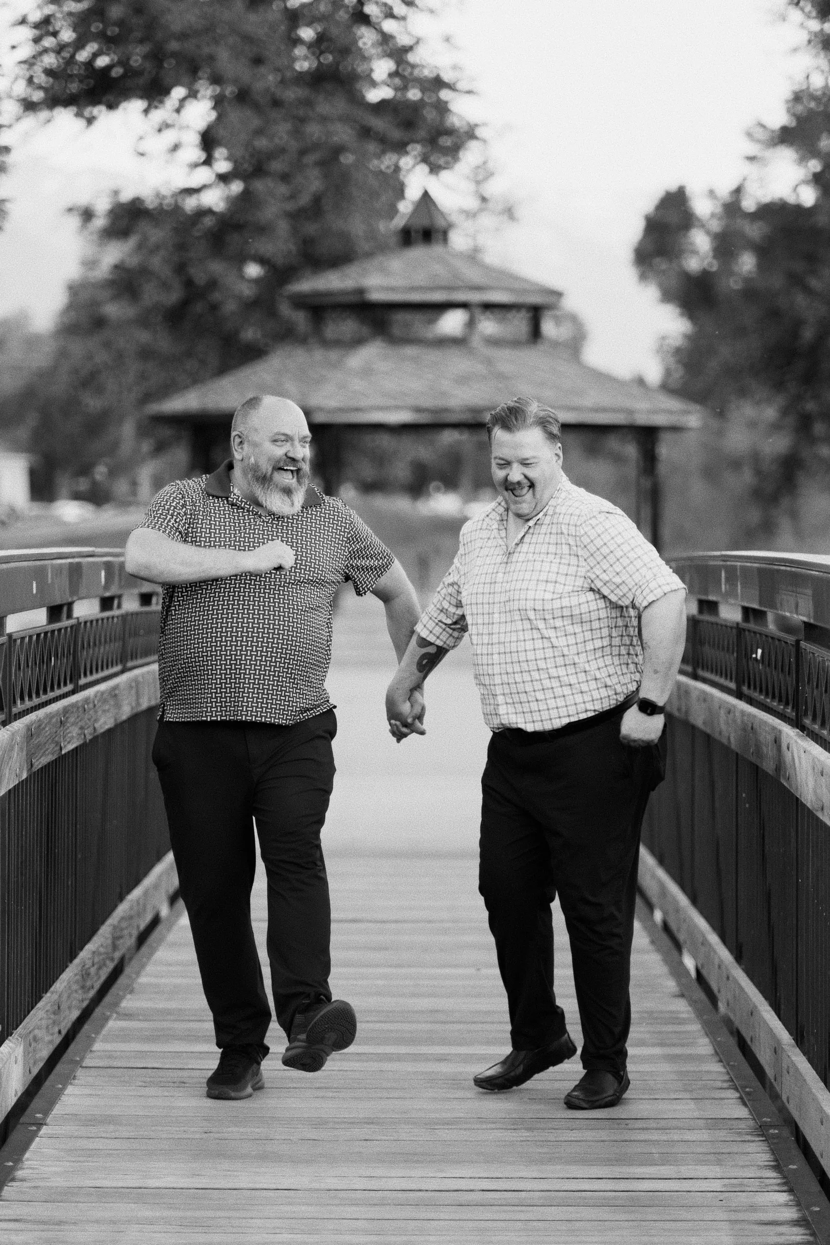 Black-and-white photo of two men laughing as they walk across a wooden footbridge.