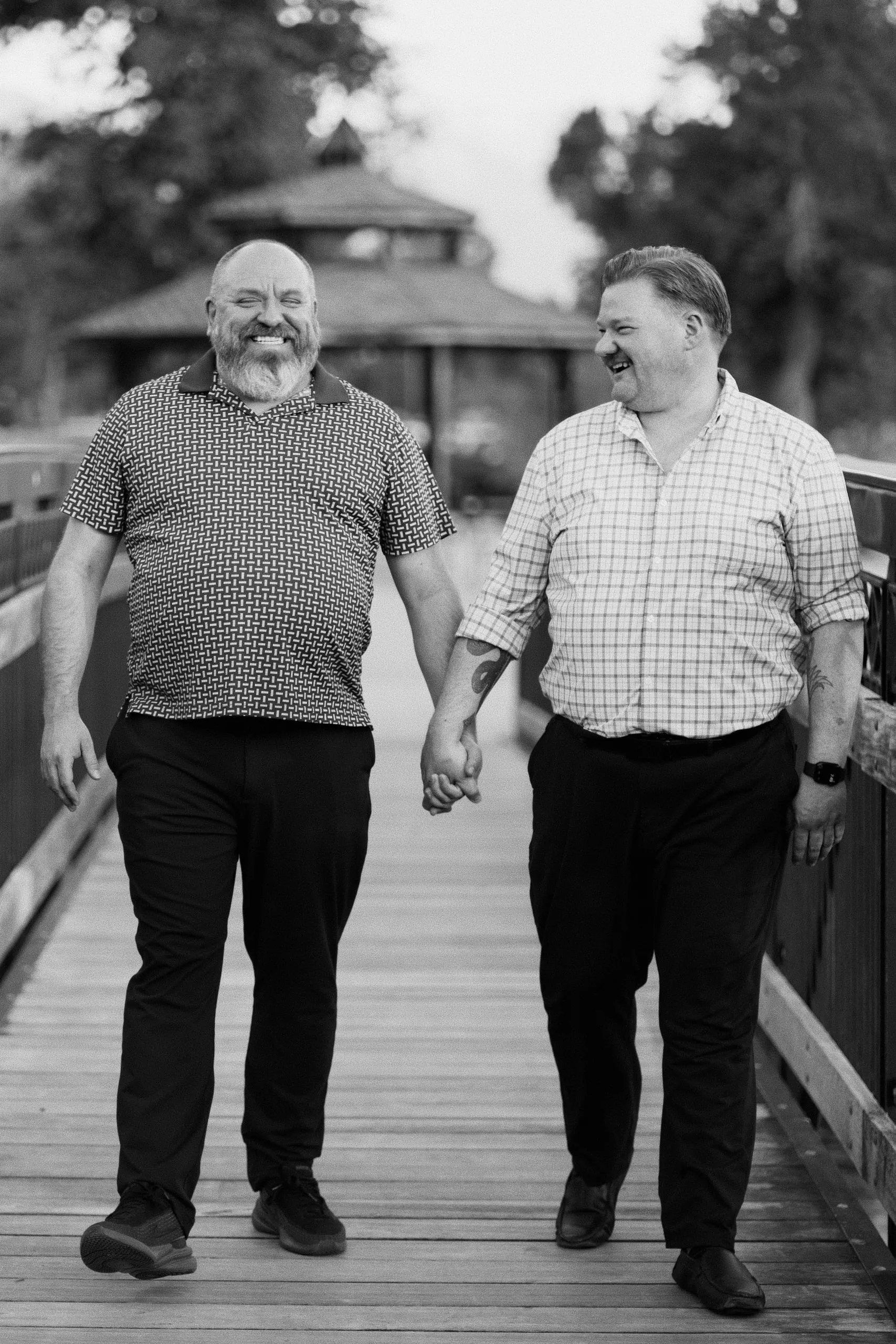 Black-and-white photo of two men holding hands as they walk toward the camera across a wooden bridge.