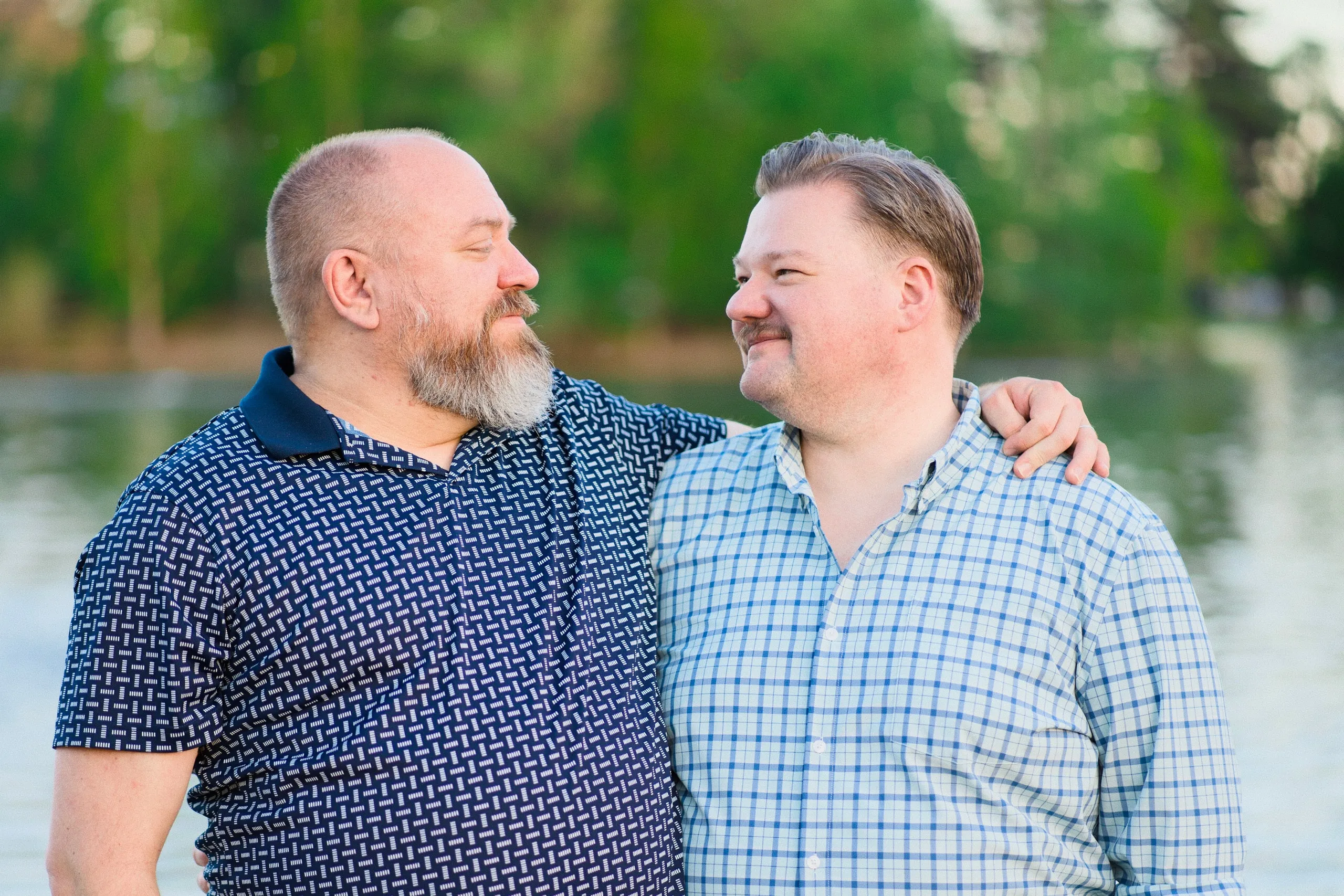 Two men stand arm in arm by a lake at golden hour, smiling at each other.