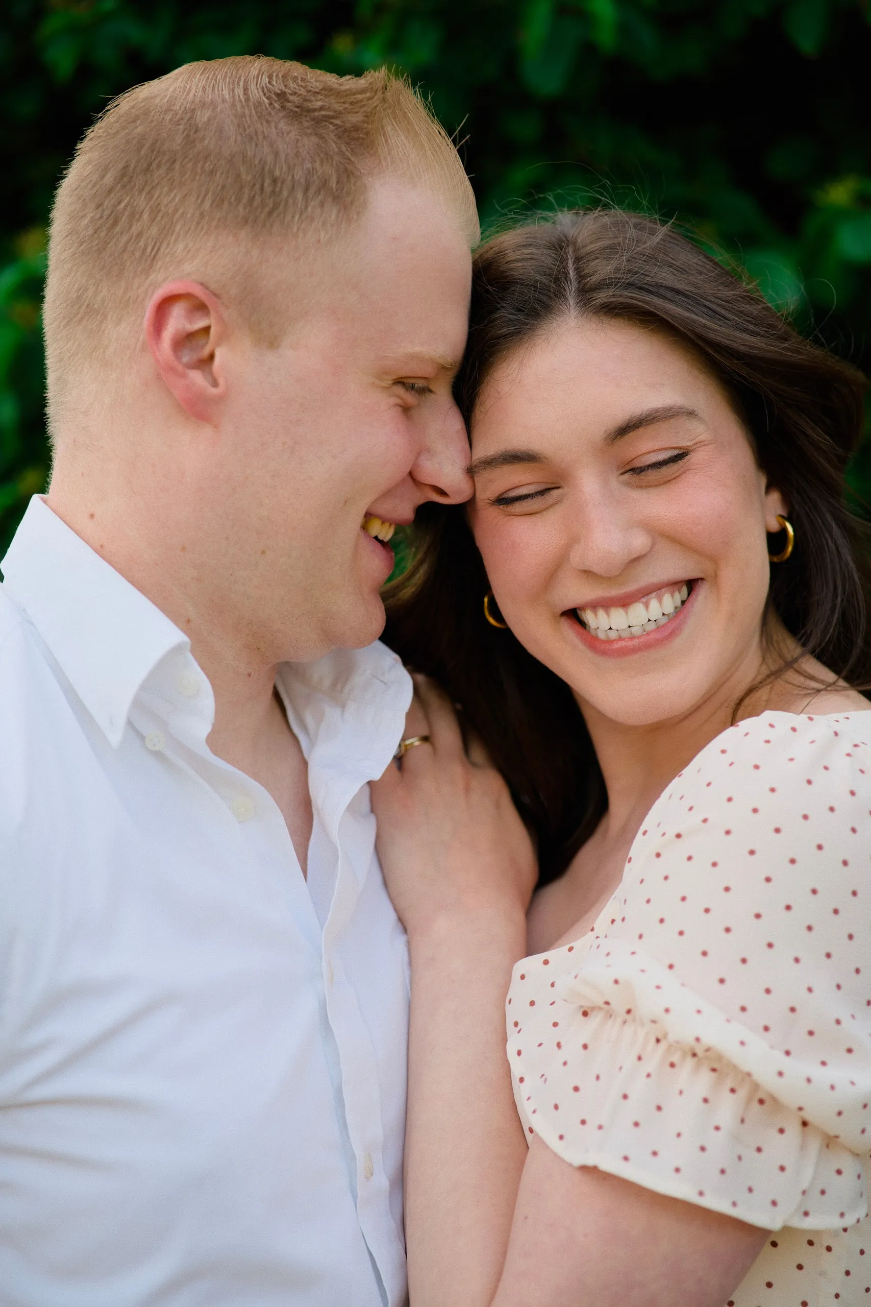 Close-up of a couple laughing with their foreheads together, the woman in a polka-dot dress, lush greenery behind.