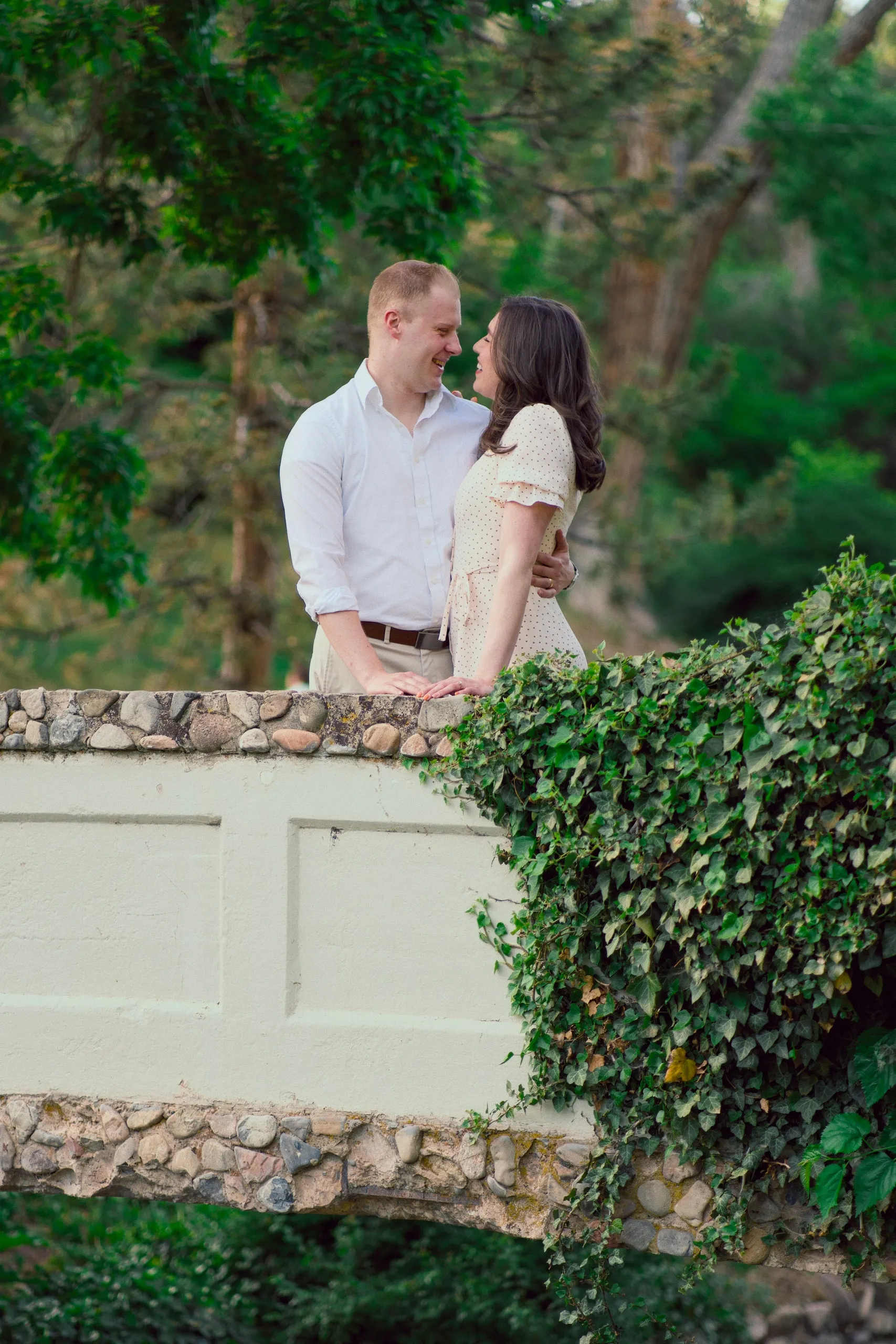 A couple embrace on an ivy-draped stone bridge surrounded by green trees.