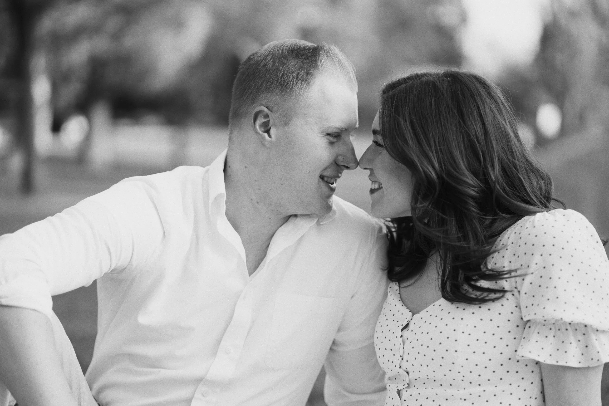 Black-and-white photo of a couple leaning in with their foreheads together, smiling, in a park.