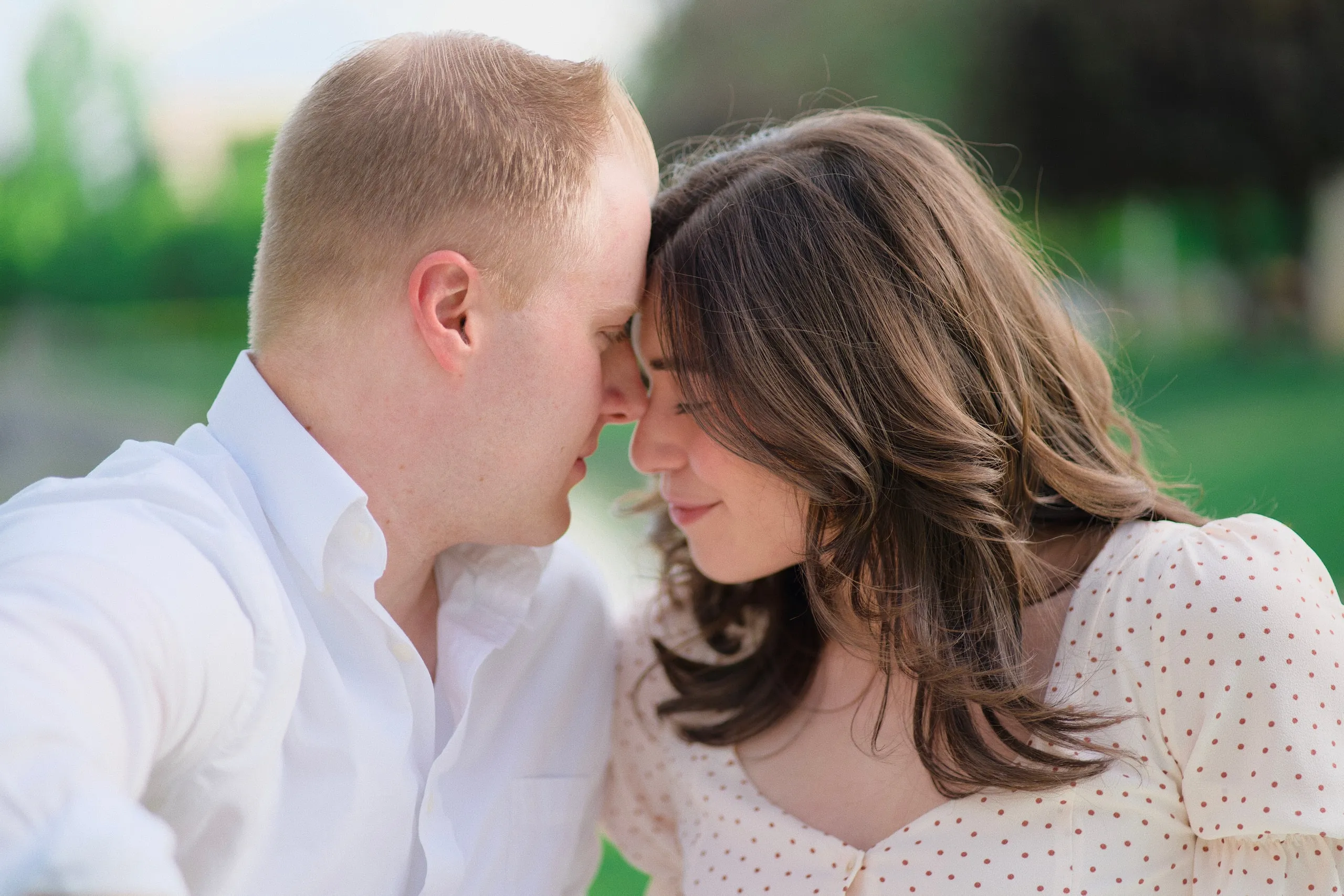 A couple rest their foreheads together with eyes closed in a sunlit park.