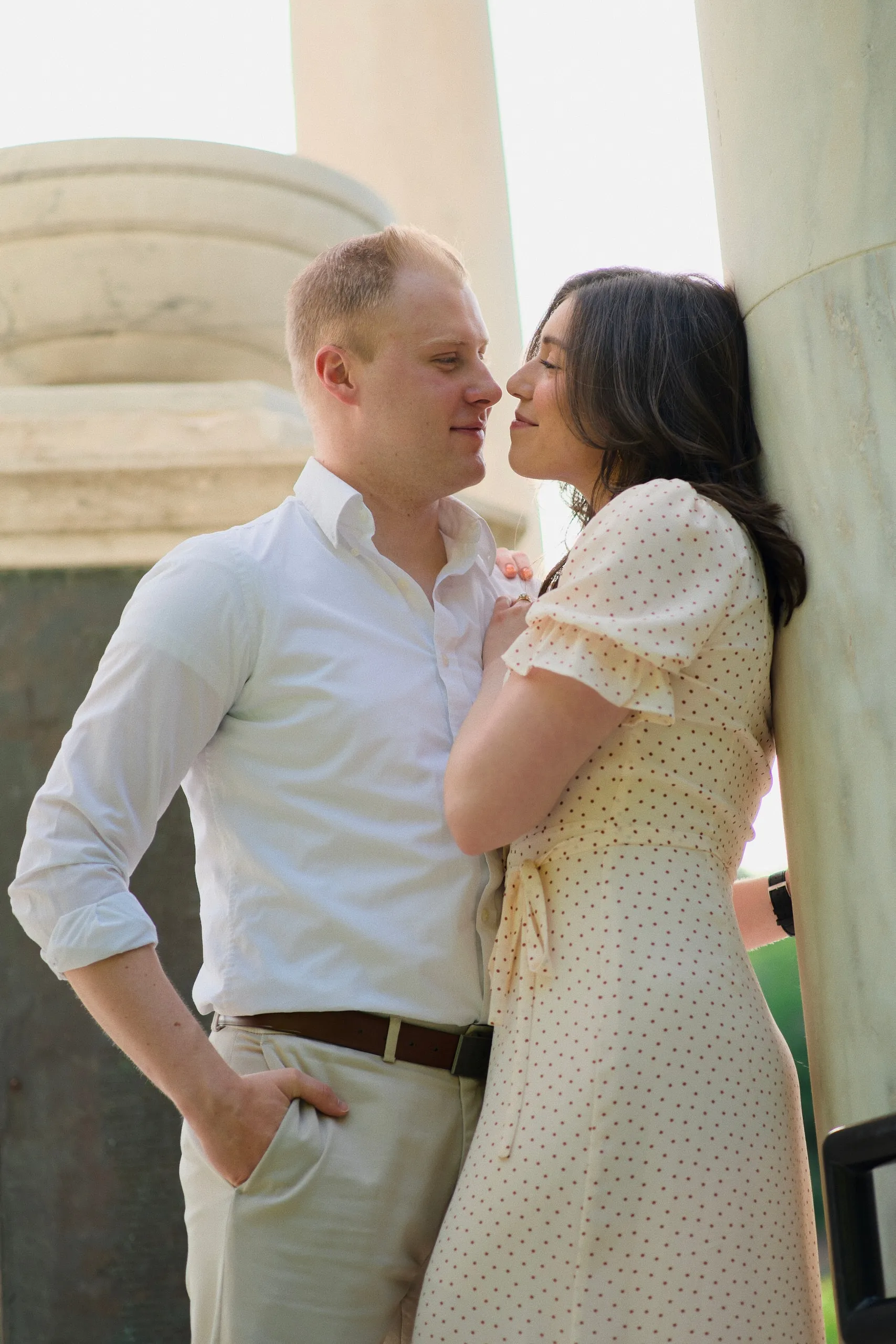 A couple lean close together among marble columns, the woman in a polka-dot dress.