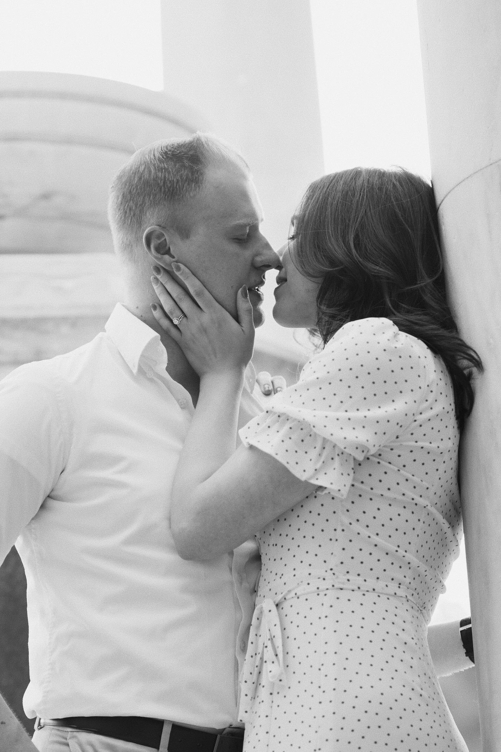 Black-and-white photo of a couple kissing against a marble column, the woman cupping the man's face.