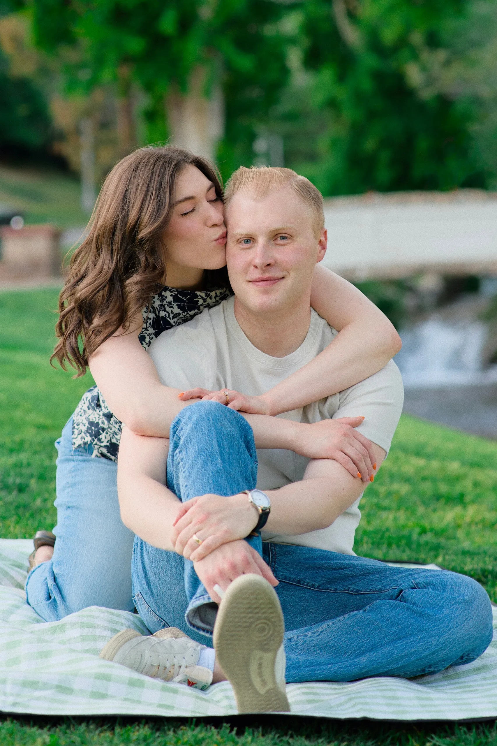 A couple sit on a picnic blanket by a waterfall as the woman kisses the smiling man's cheek.