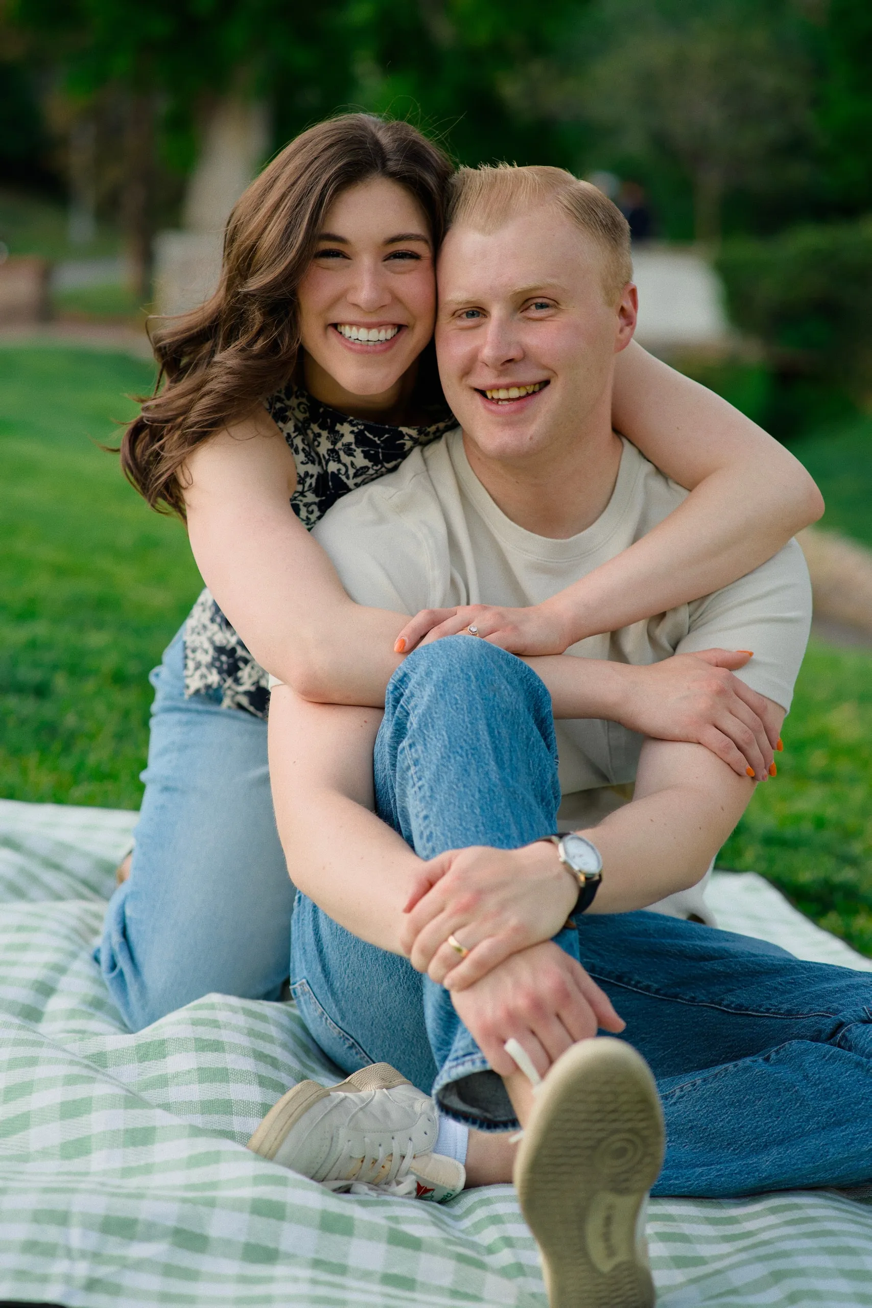 A couple sit on a picnic blanket in a park, the woman hugging the man as they both laugh.