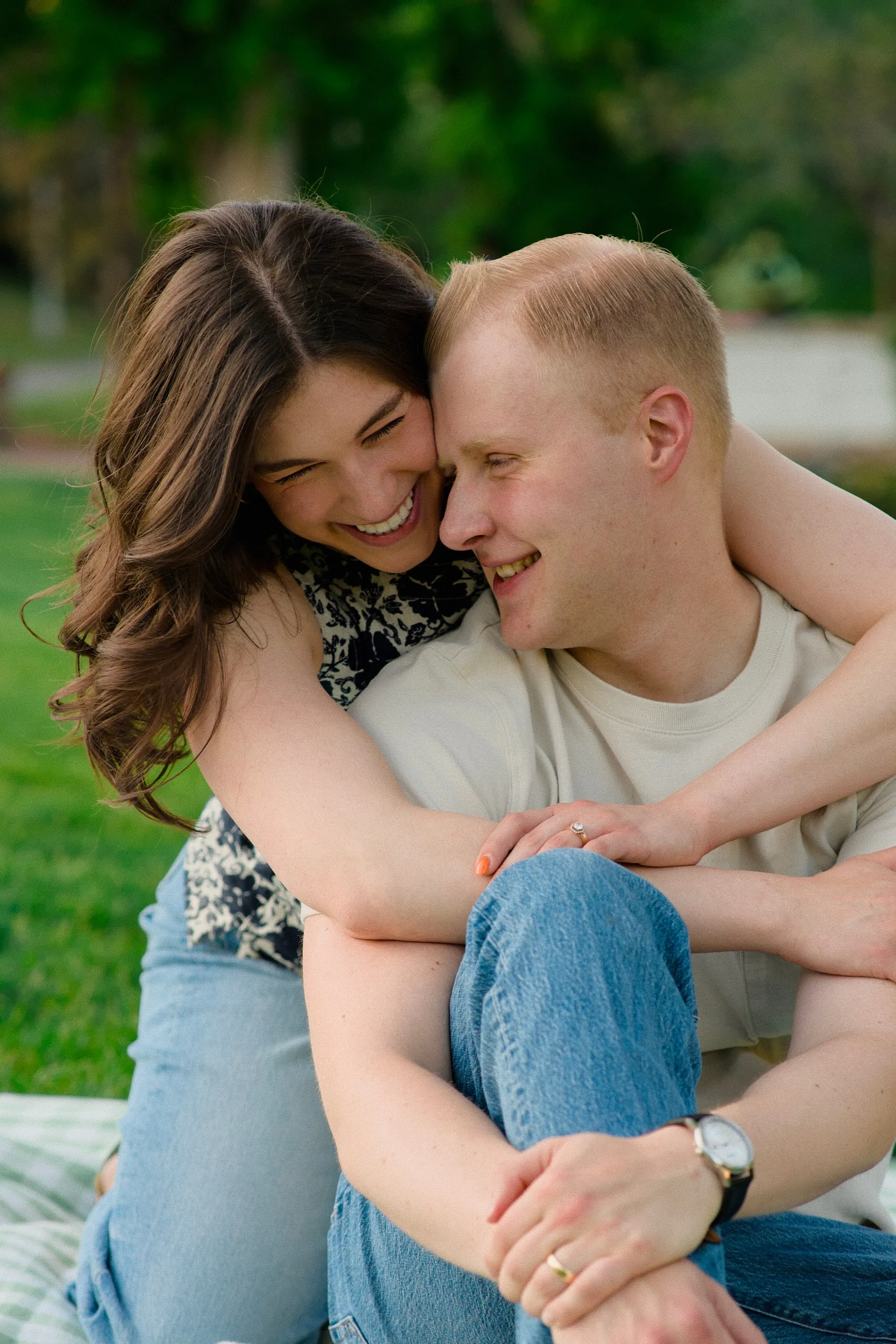 A couple laugh together on the grass, the woman nuzzling into the man's shoulder.