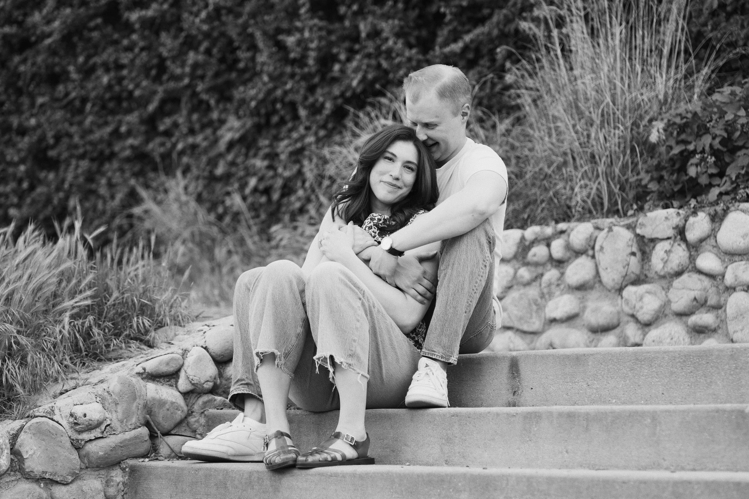 Black-and-white photo of a couple sitting close on stone steps as the man kisses the top of the woman’s head.