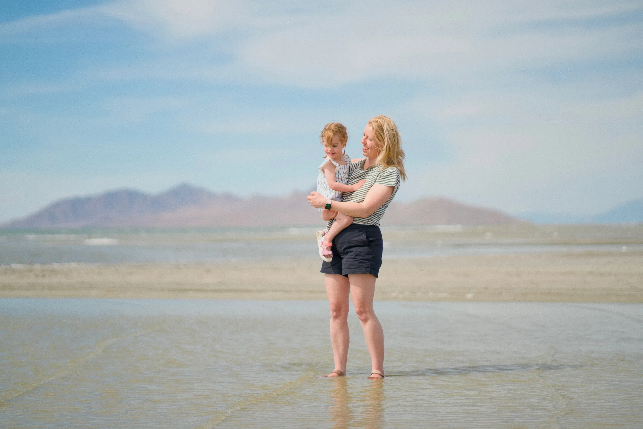 A woman stands barefoot in the shallow lake water holding her toddler daughter, distant mountains behind.