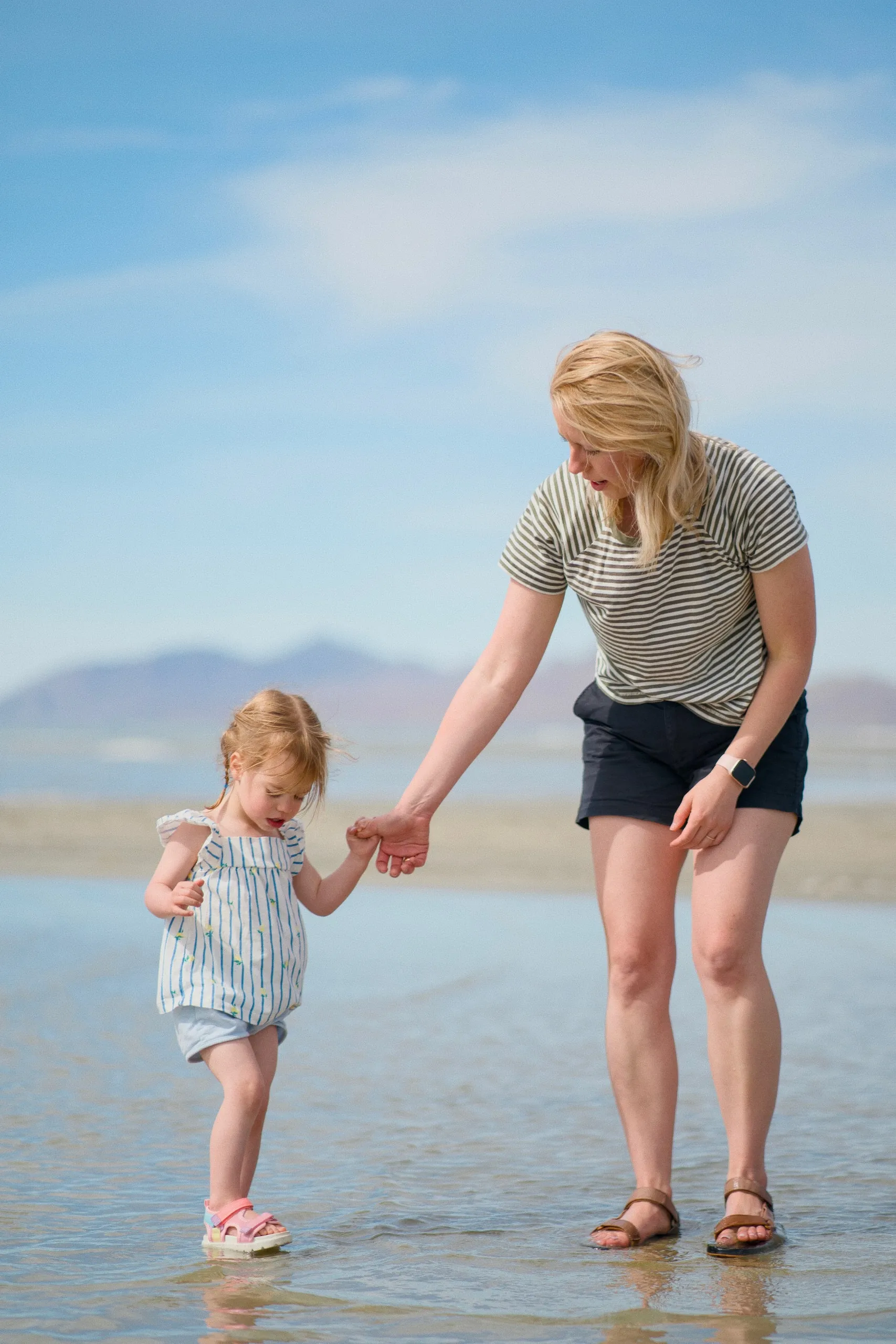 A mother holds her toddler daughter's hand as the little girl wades in the shallow lake water.