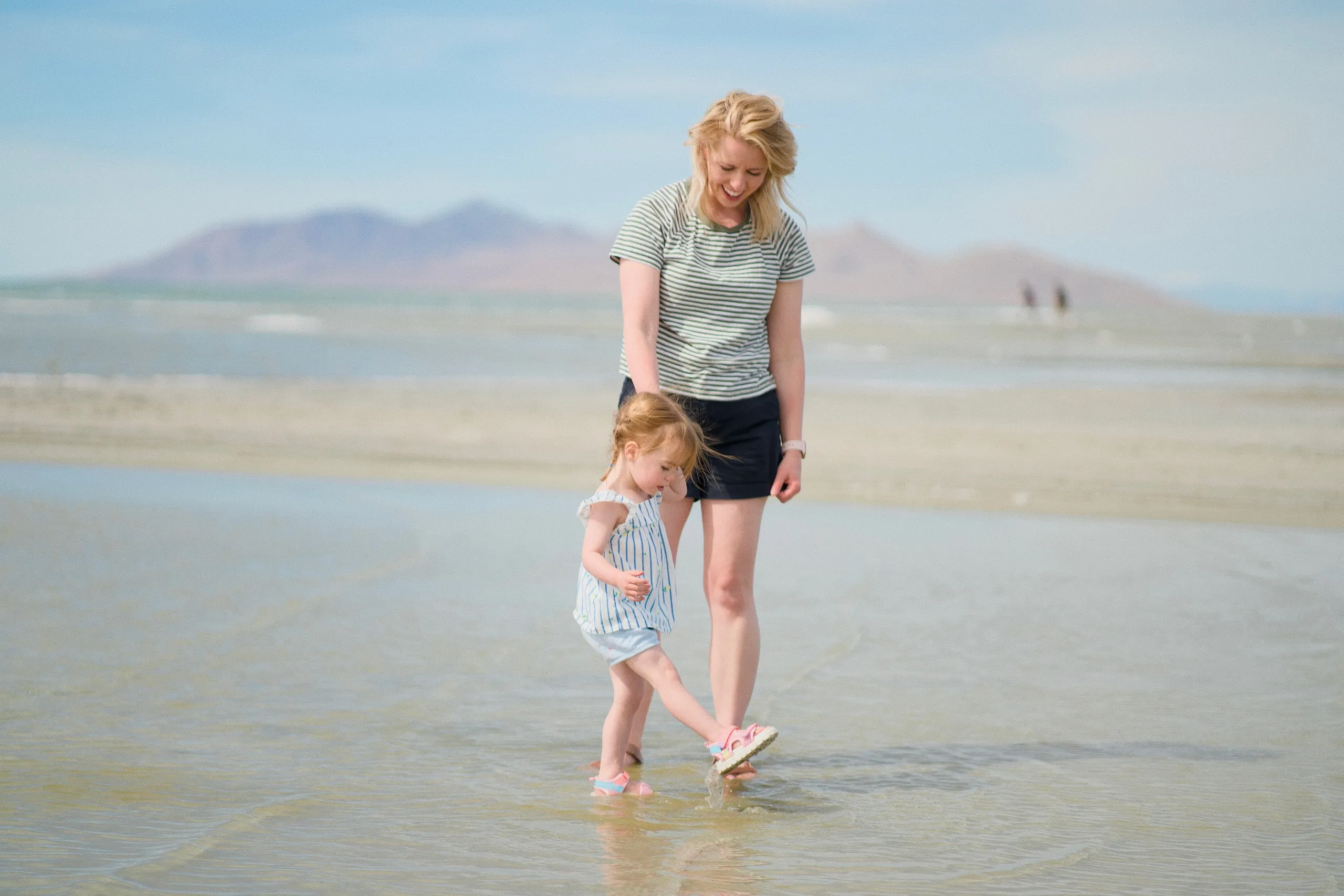 A mother and her toddler daughter wade together at the lake's edge, looking down at the water.
