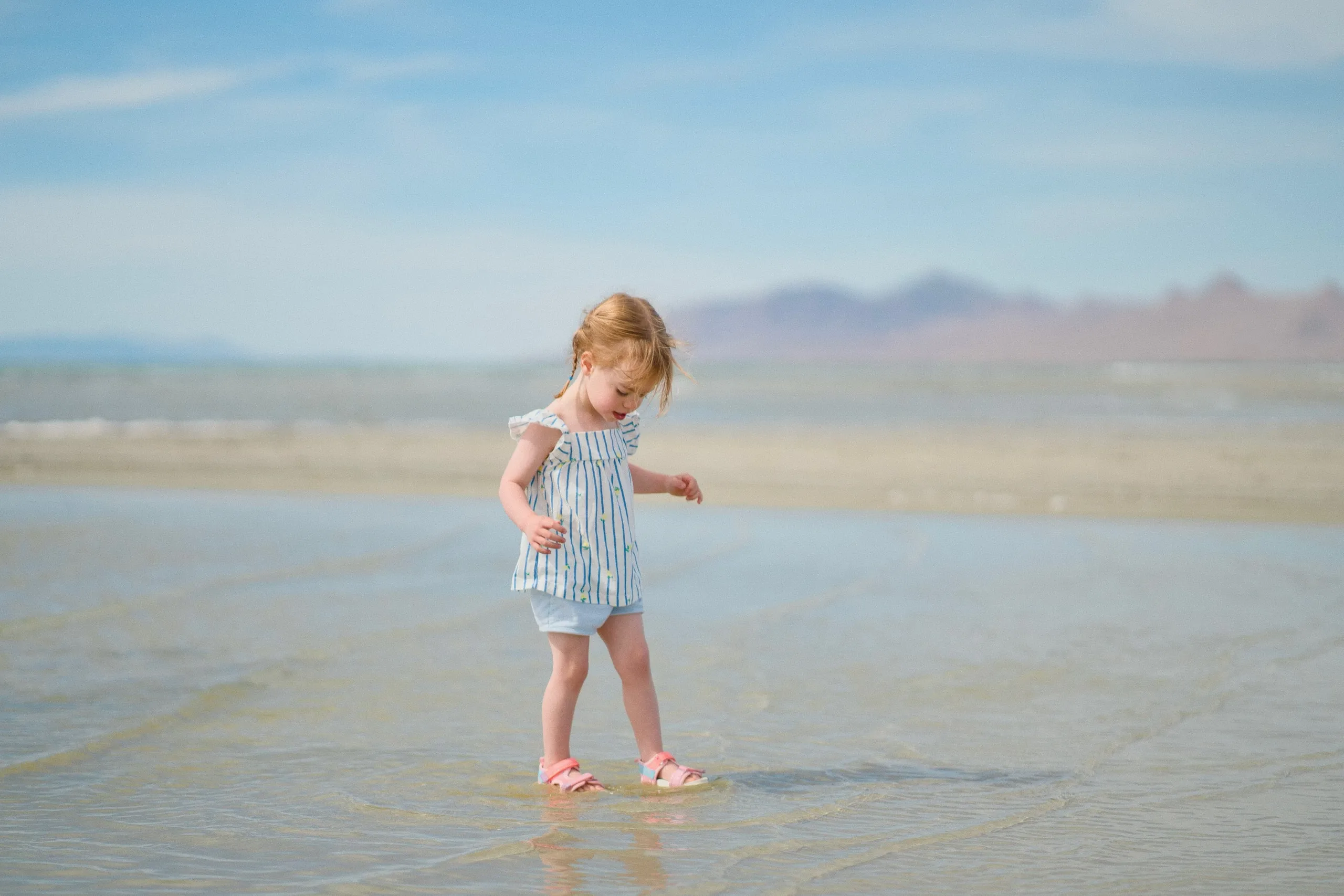 A toddler girl in a striped dress plays in the shallow lake water, mountains in the distance.