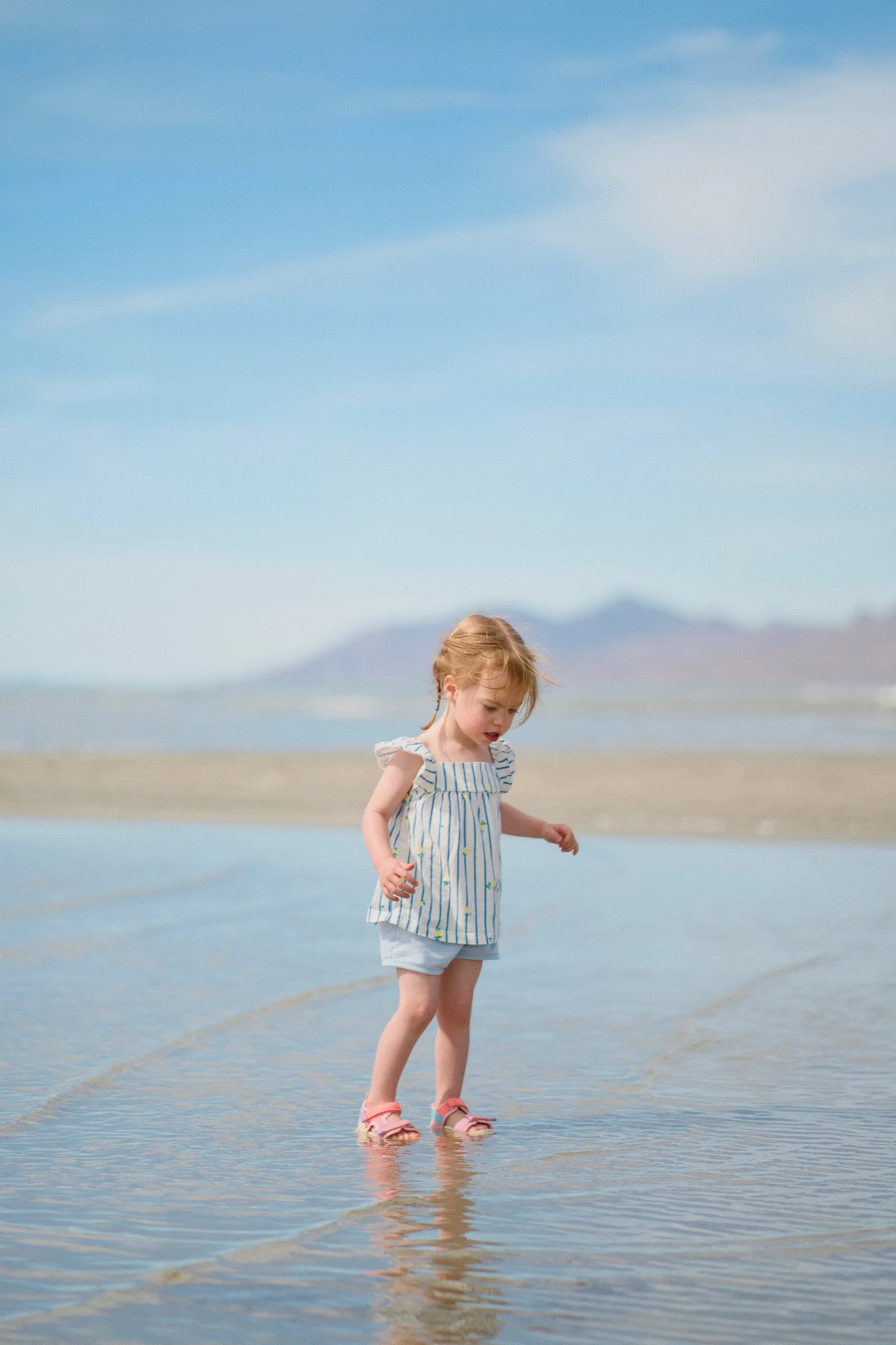 A little girl in a striped dress walks through the shallow lake water, looking down.