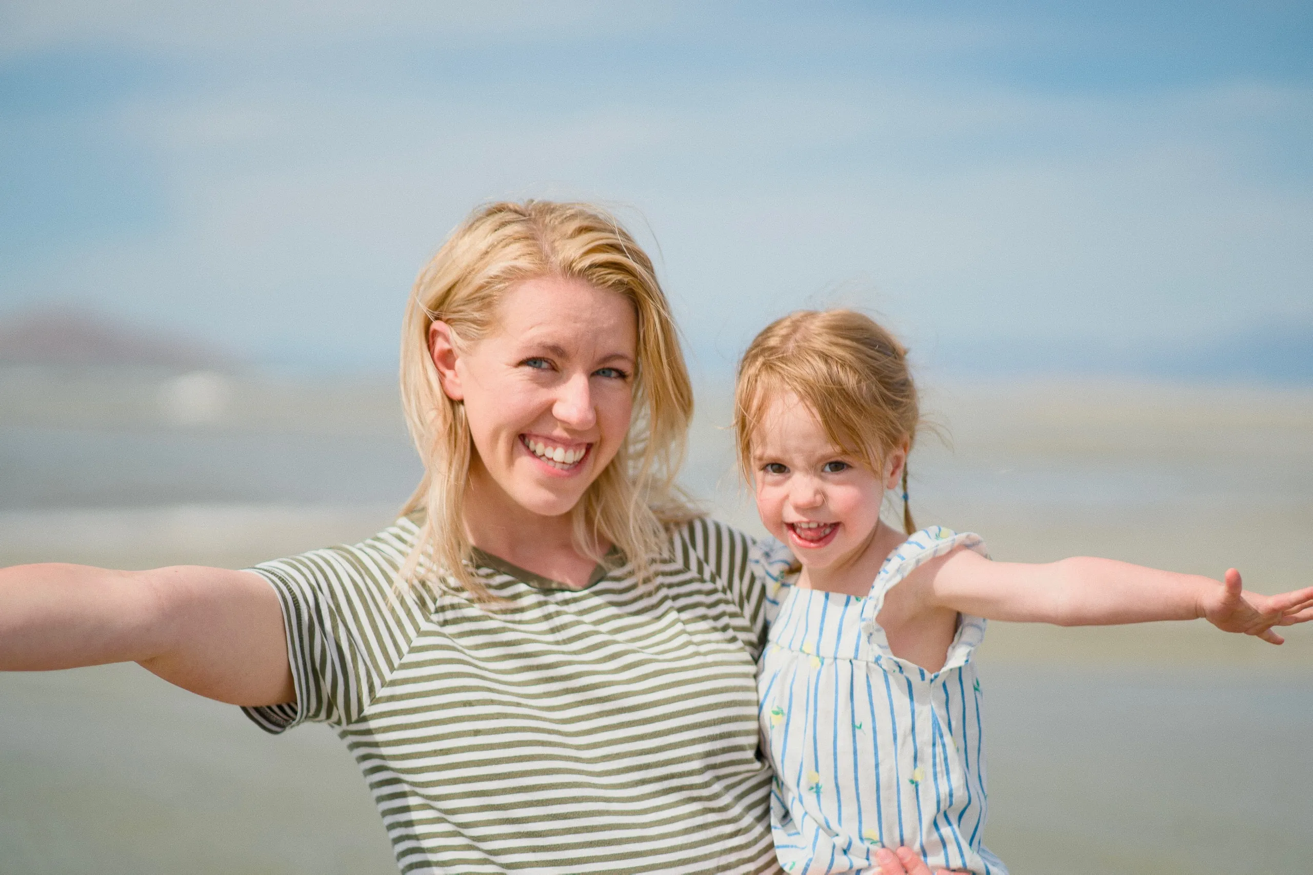 A blonde woman and her young daughter smile at the camera with their arms outstretched by the lake.