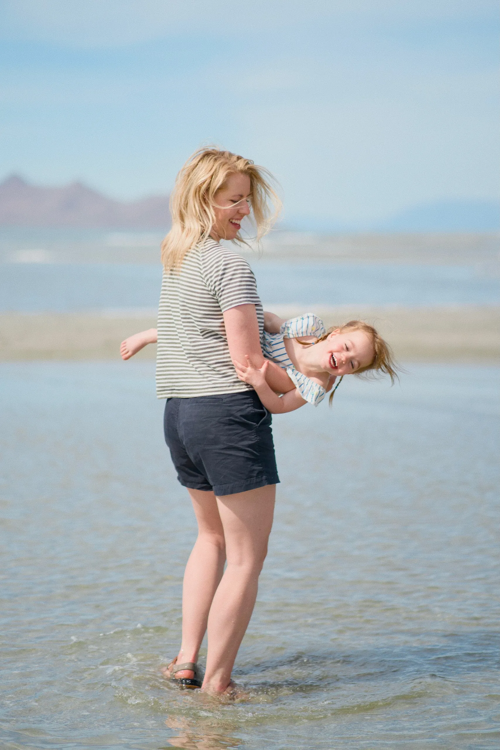 A mother playfully swings her laughing daughter while standing in the shallow lake water.