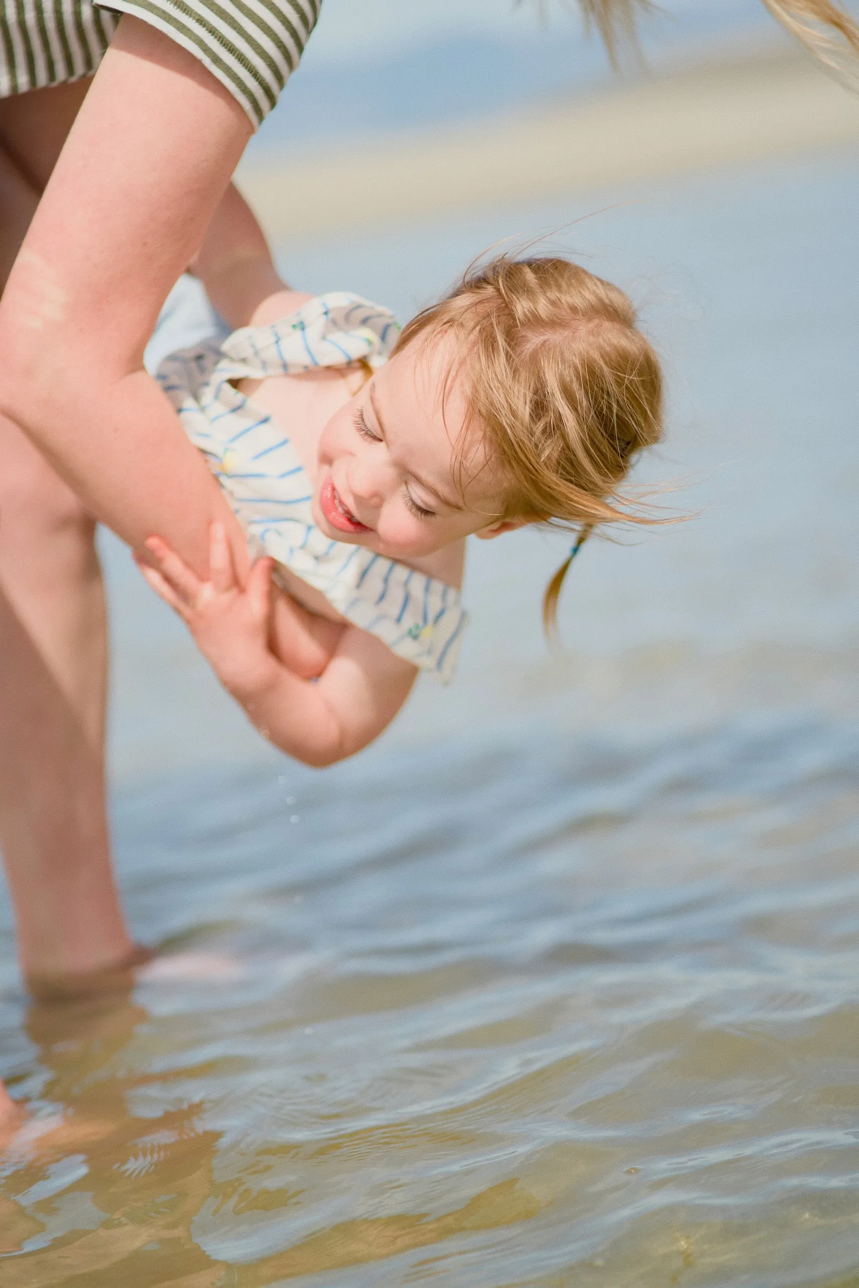 A mother holds her giggling toddler daughter over the shallow lake water.