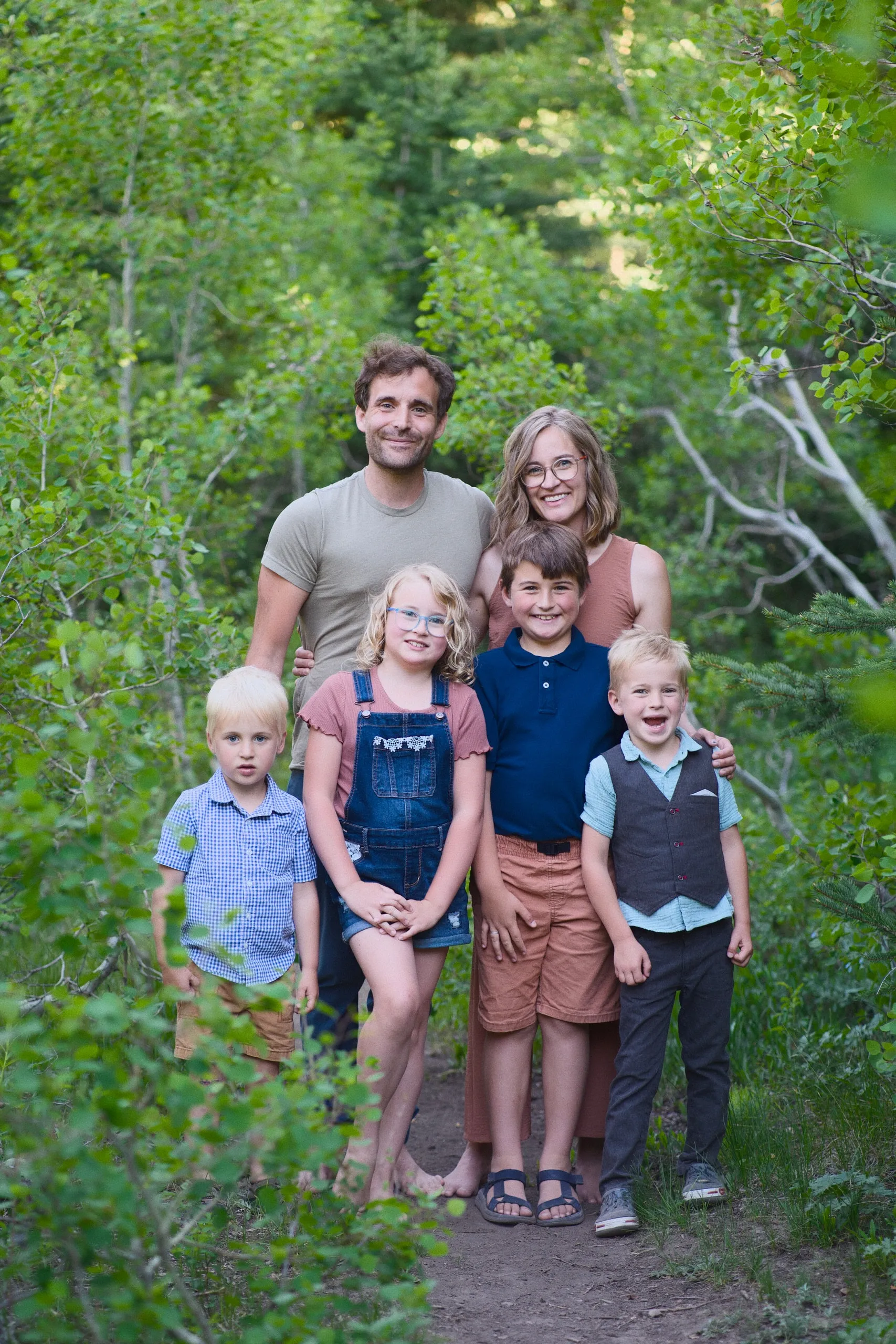 A family of six stands together on a leafy wooded trail, the four children in front of their smiling parents.