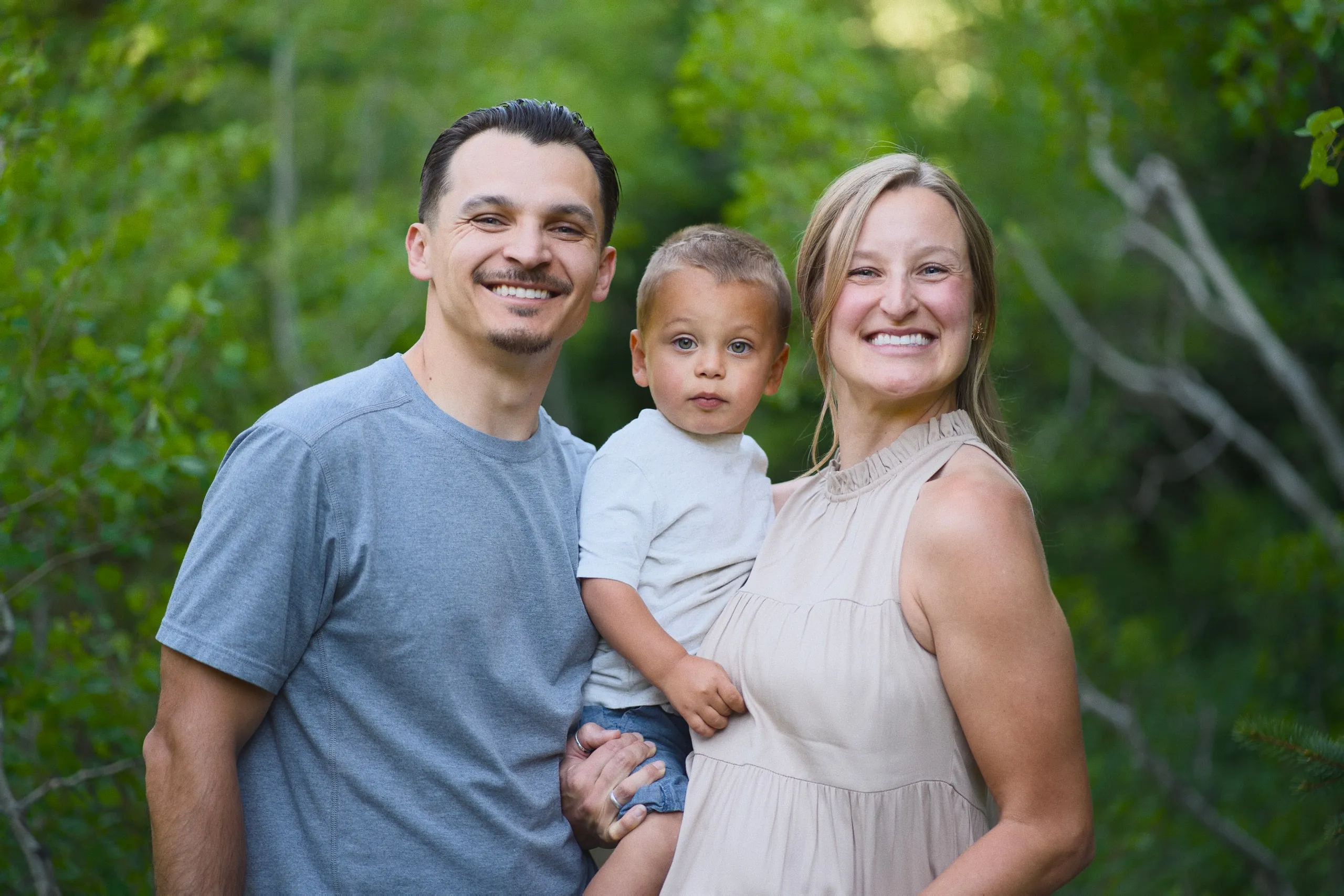 A couple hold their young son between them, all three smiling, with green foliage behind.