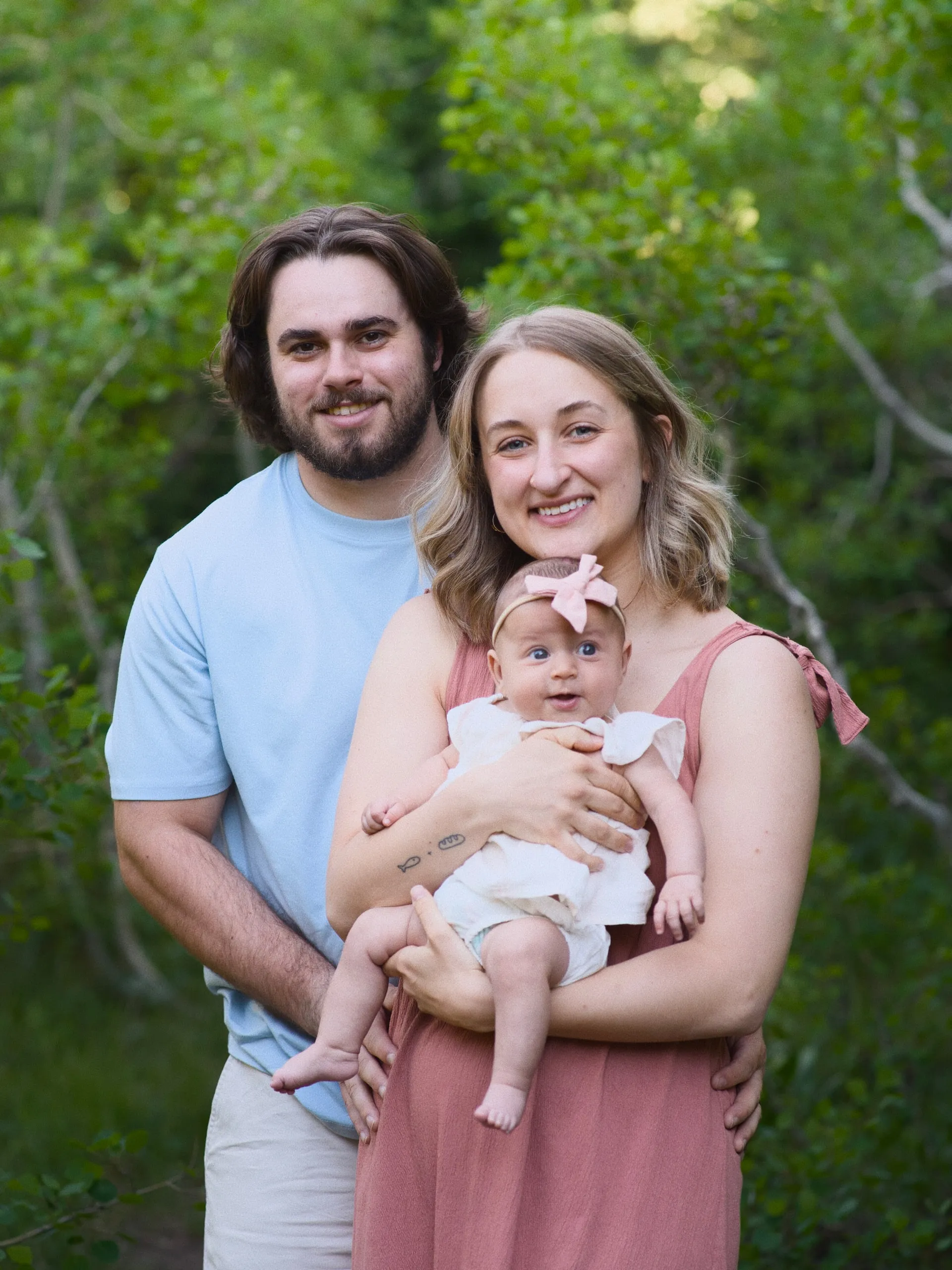A young couple cradle their baby daughter, who wears a pink bow, in front of lush green leaves.