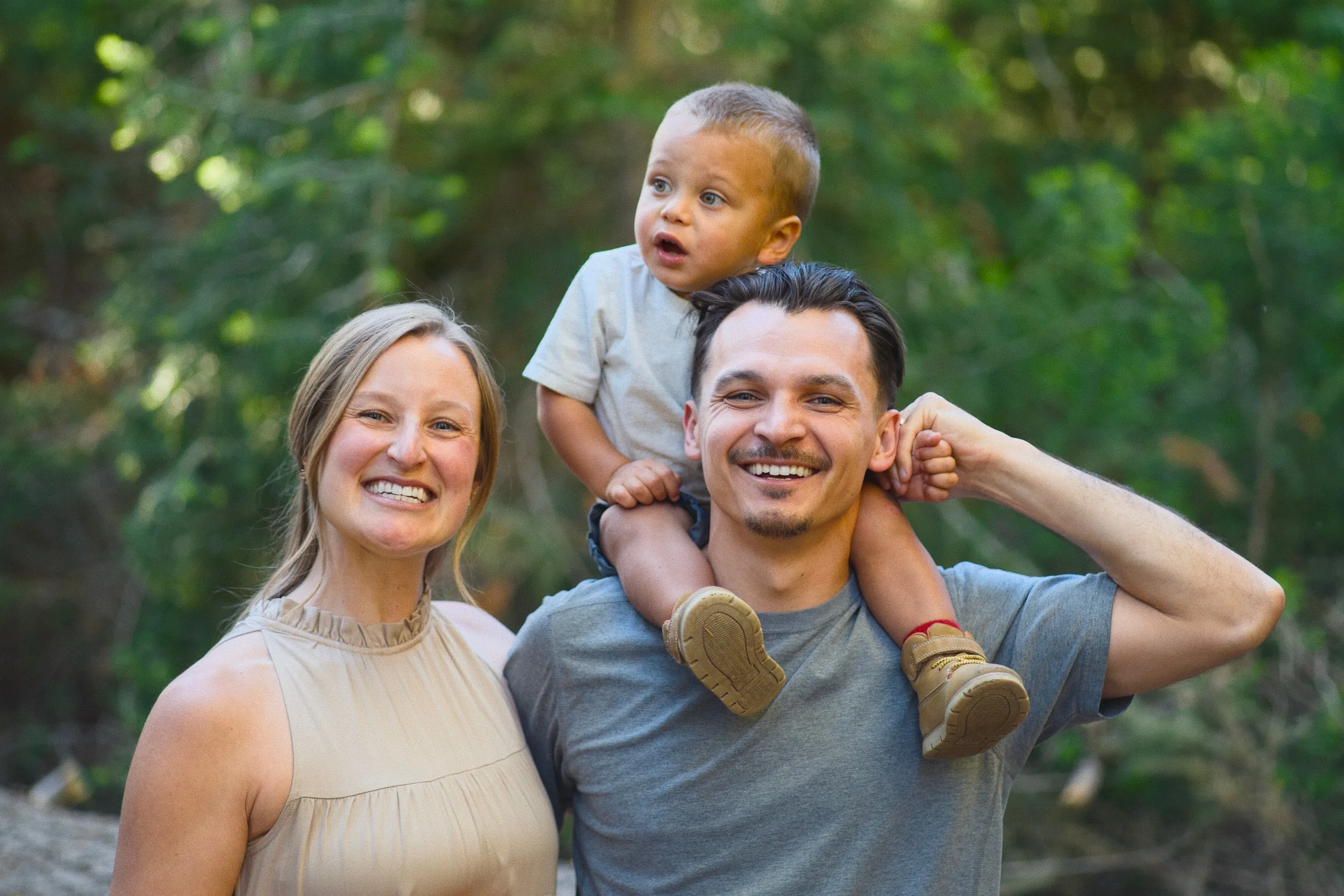 A smiling couple pose in the woods with their young son riding on the father’s shoulders.