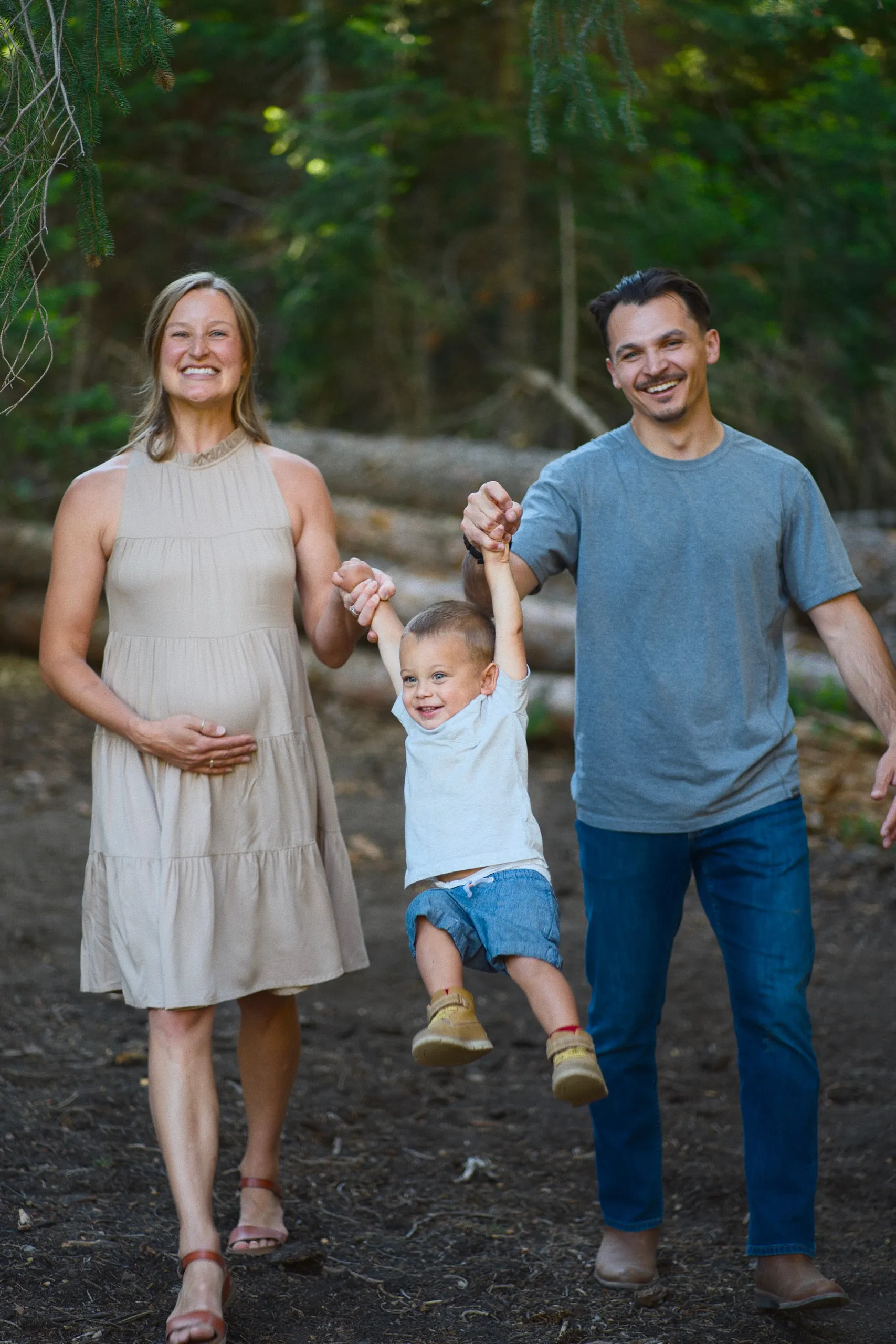 An expecting mother and a father swing their giggling young son by the arms as they walk through the woods.