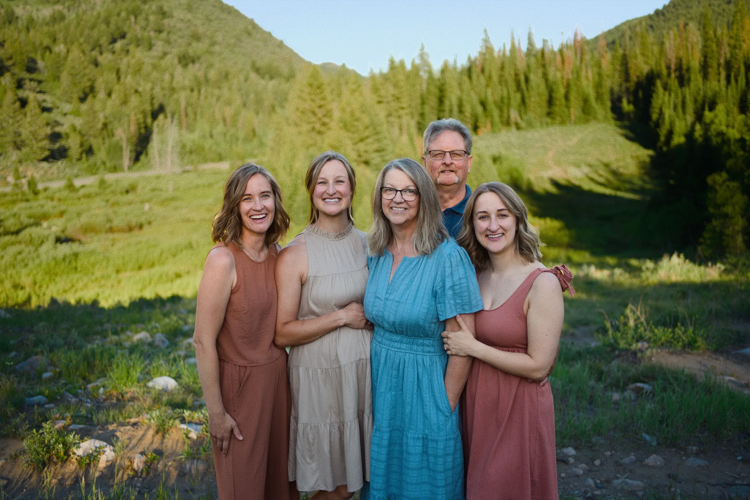 Five adults stand arm in arm in a green mountain meadow at golden hour.