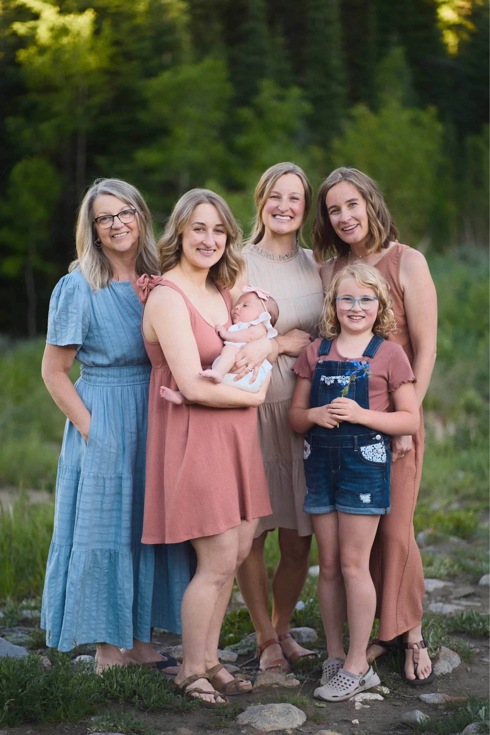 A group of women and girls across generations gather in a mountain meadow, one cradling a swaddled baby.
