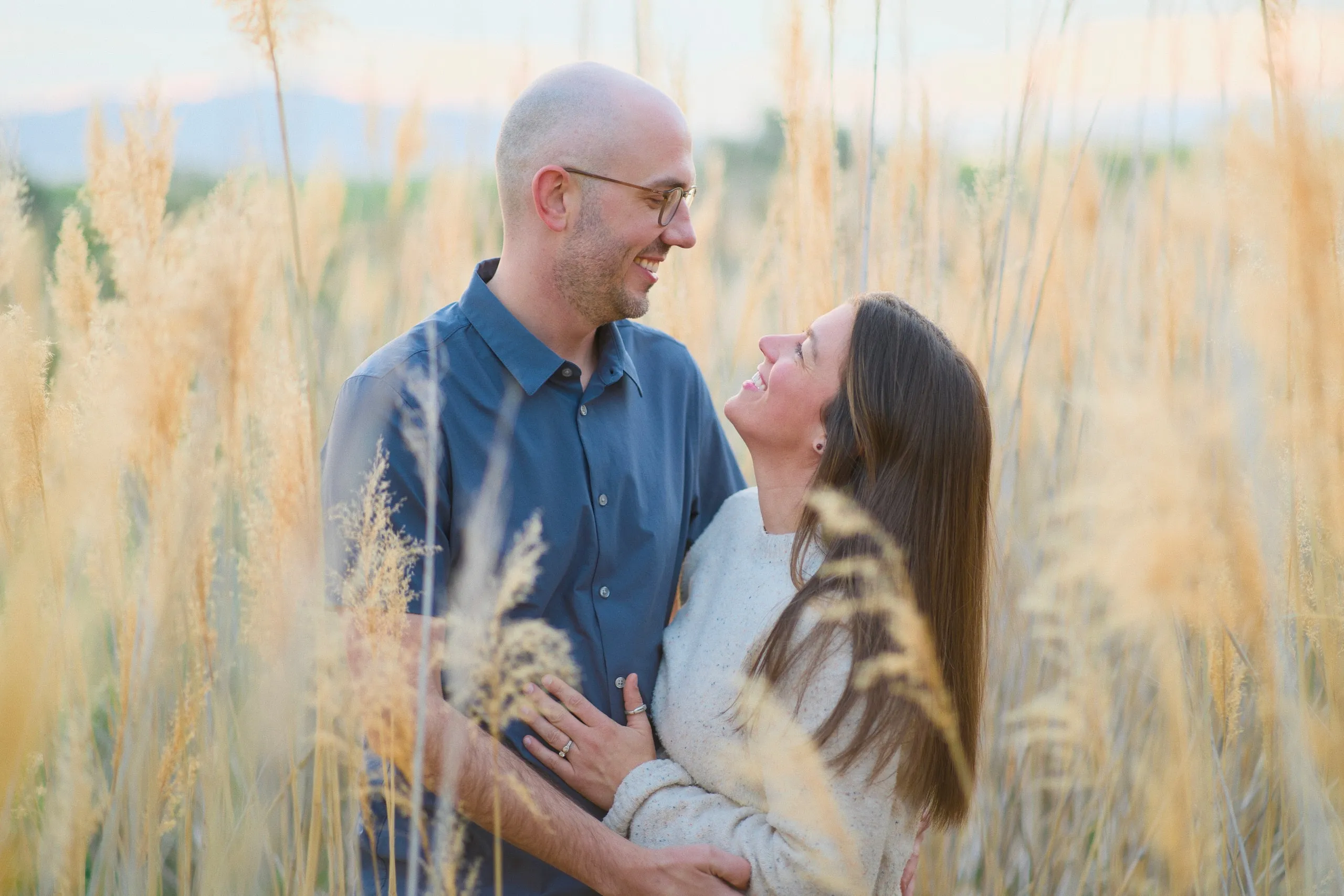 A couple stand face to face in a field of tall golden grasses at sunset.